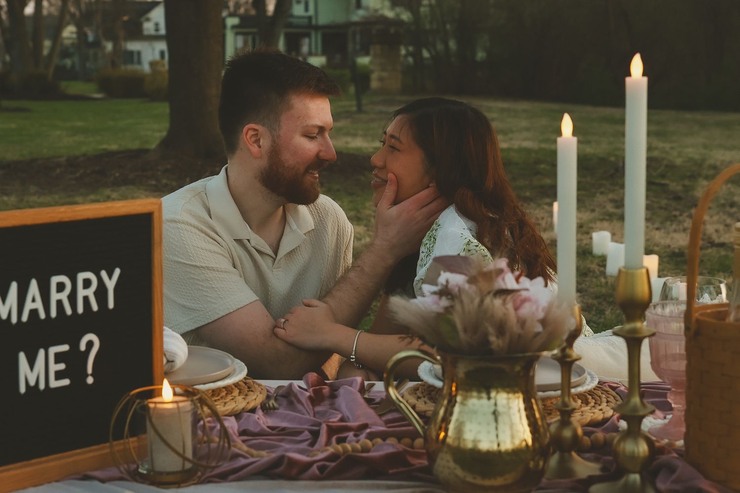 Romantic outdoor candlelit dinner proposal setup with chalkboard sign reading 'Marry Me?' and couple sharing intimate moment.