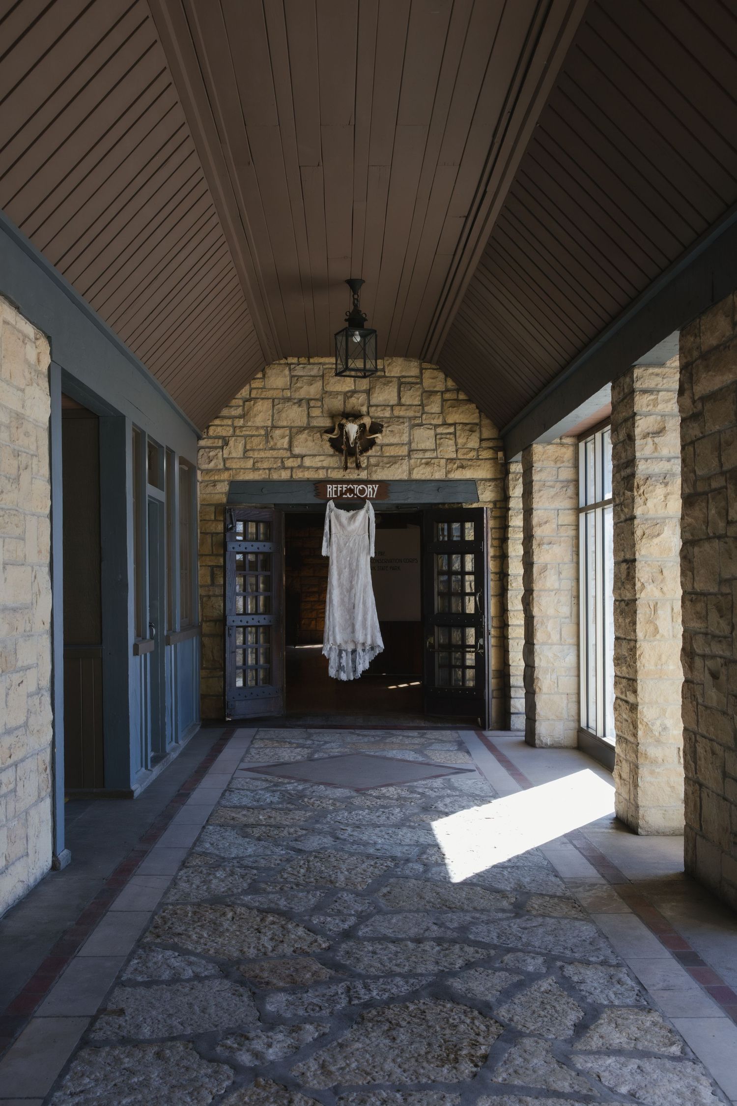A white wedding dress hangs in a stone-walled breezeway with blue doors and natural light streaming through windows.