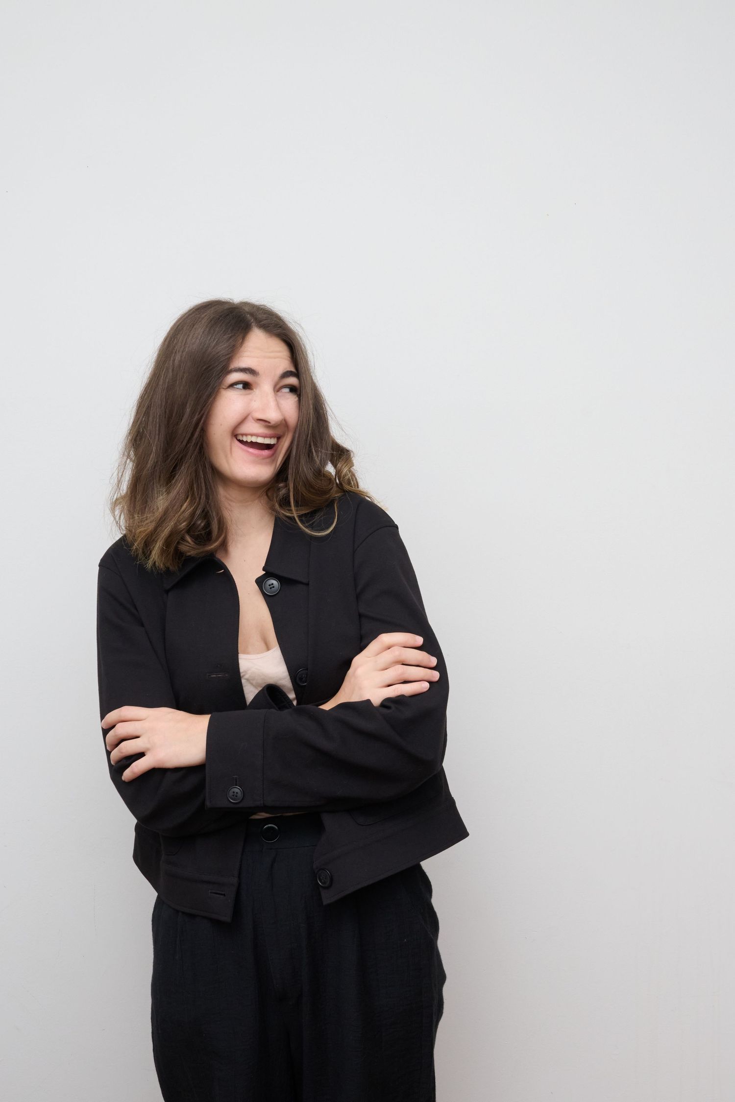 Person in all black outfit laughing with arms crossed against white background.
