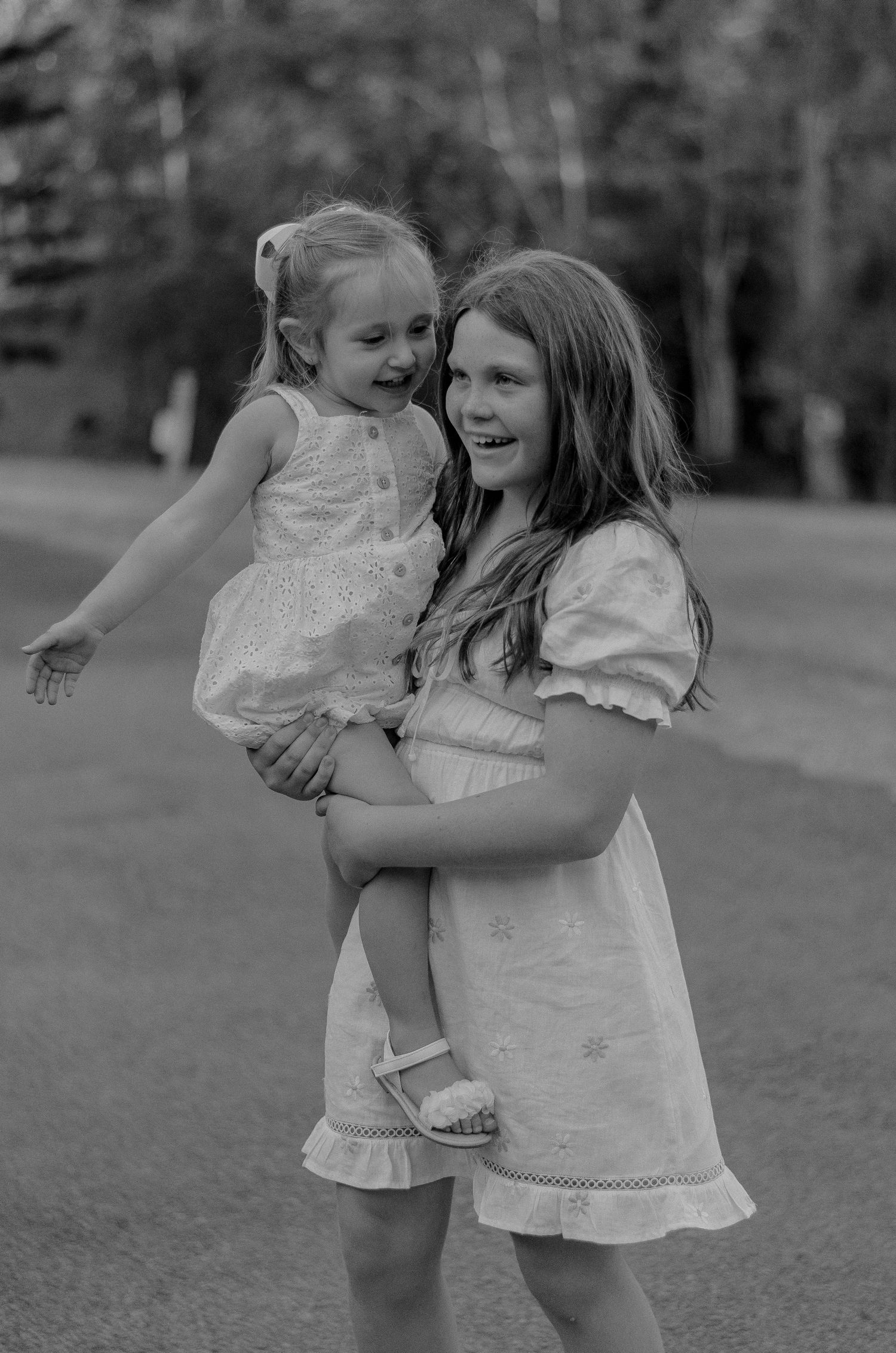 Two children in white dresses playing and spinning together outdoors in black and white photography.