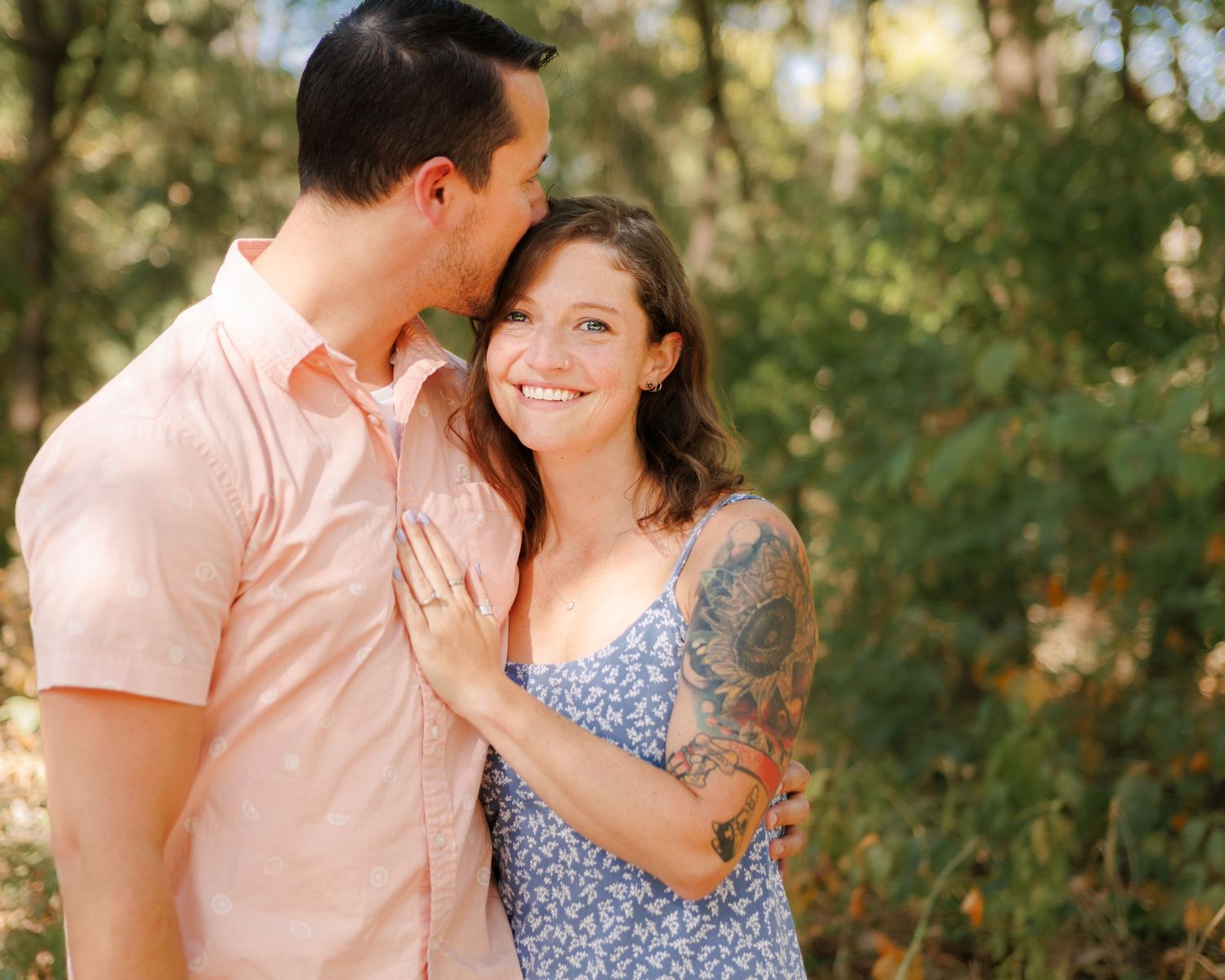 A joyful couple embraces outdoors in a natural setting with soft sunlit foliage in the background.