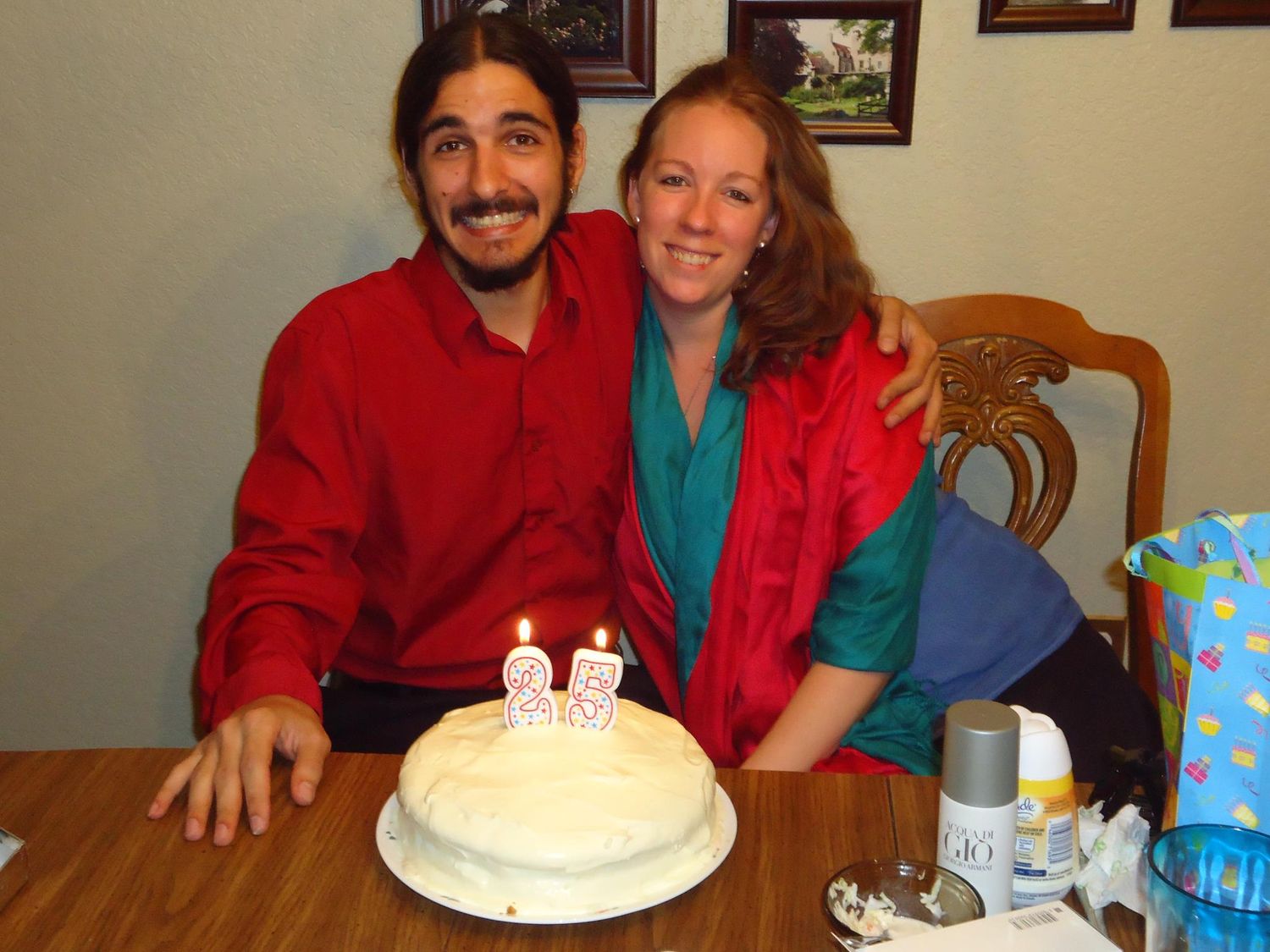 Two people sit at a table smiling with a birthday cake topped with candles, wearing red and turquoise jackets.