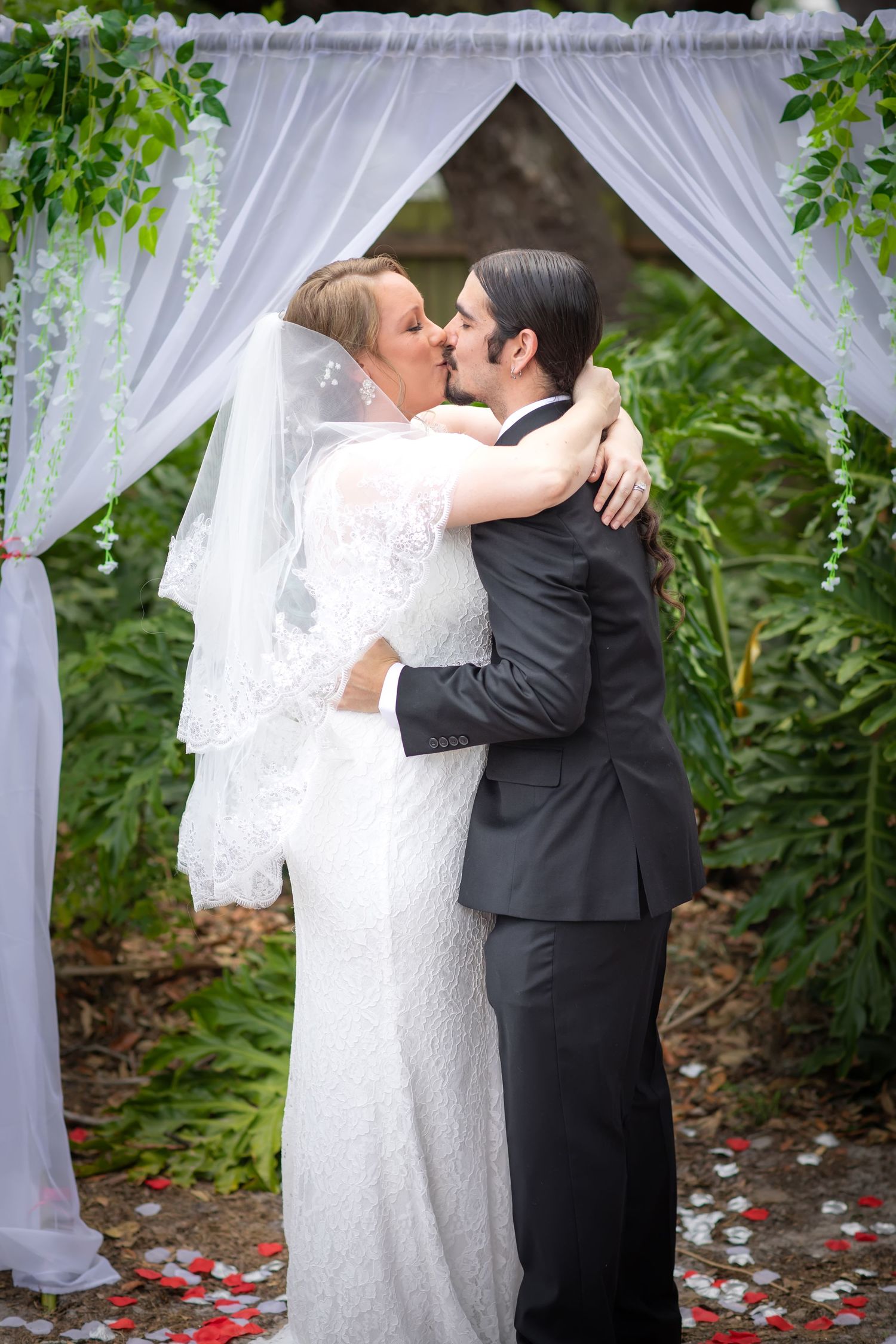 A romantic wedding scene with white curtains and flower petals under a floral arch in a garden. Tampa Photographer, Tampa Videographer,