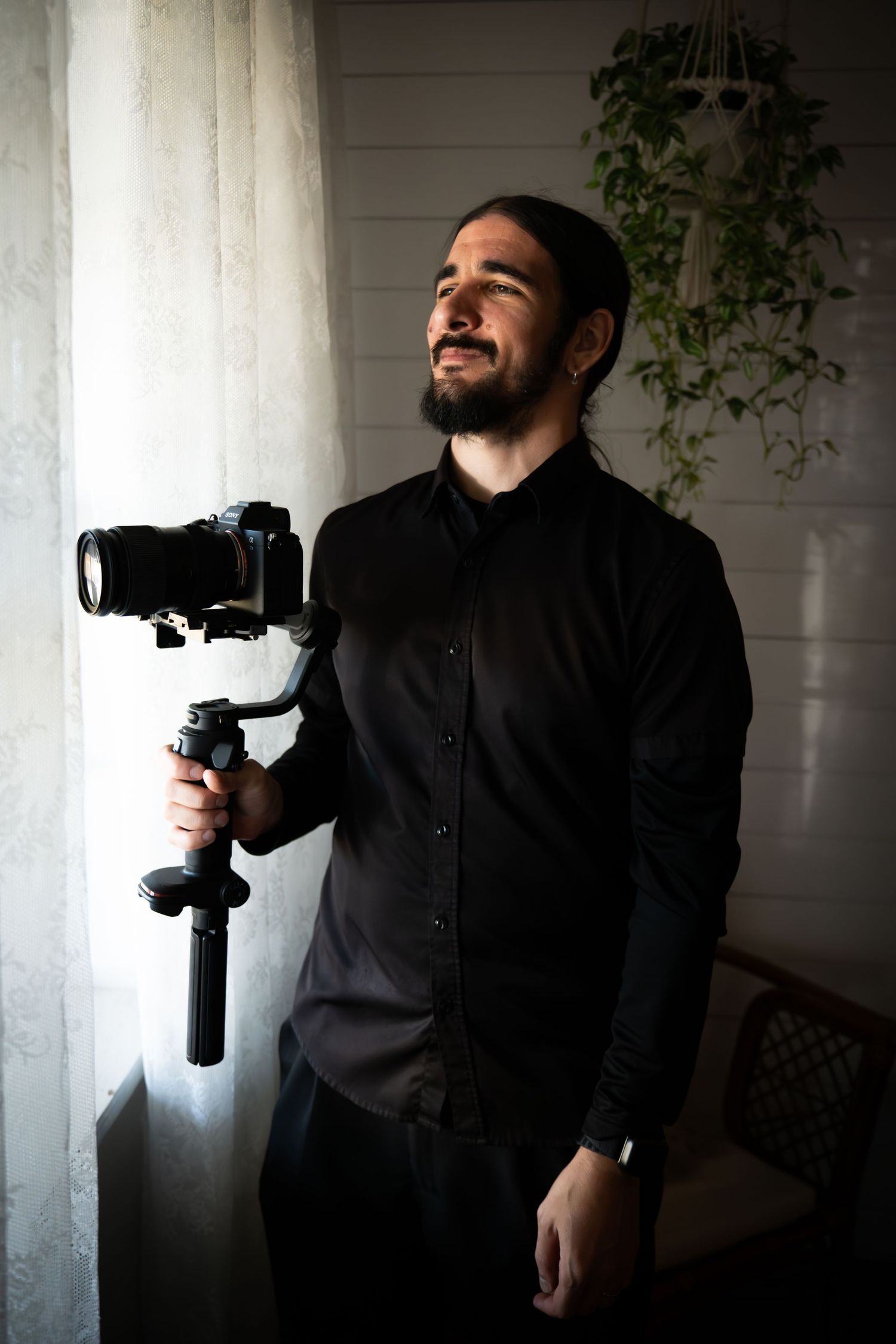 Professional photographer in black shirt holding camera gimbal stabilizer by window with natural light and indoor plant. Tampa Photographer