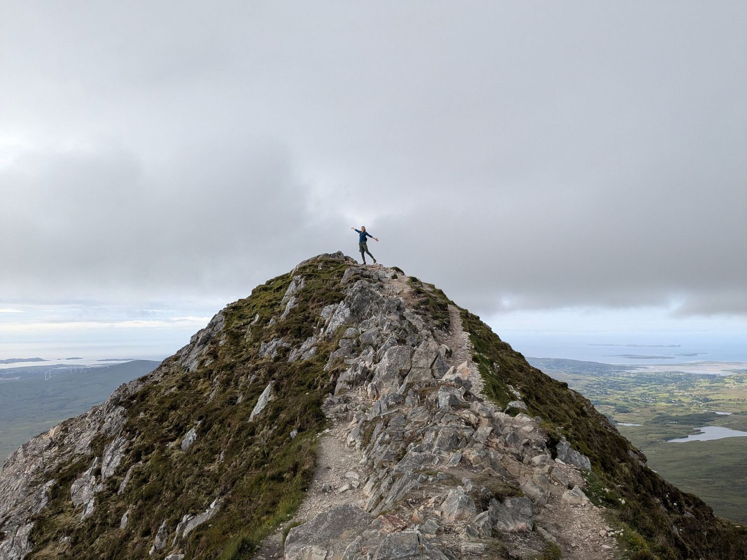 A silhouetted hiker stands triumphantly on a rocky mountain peak against a cloudy sky and sprawling landscape below.