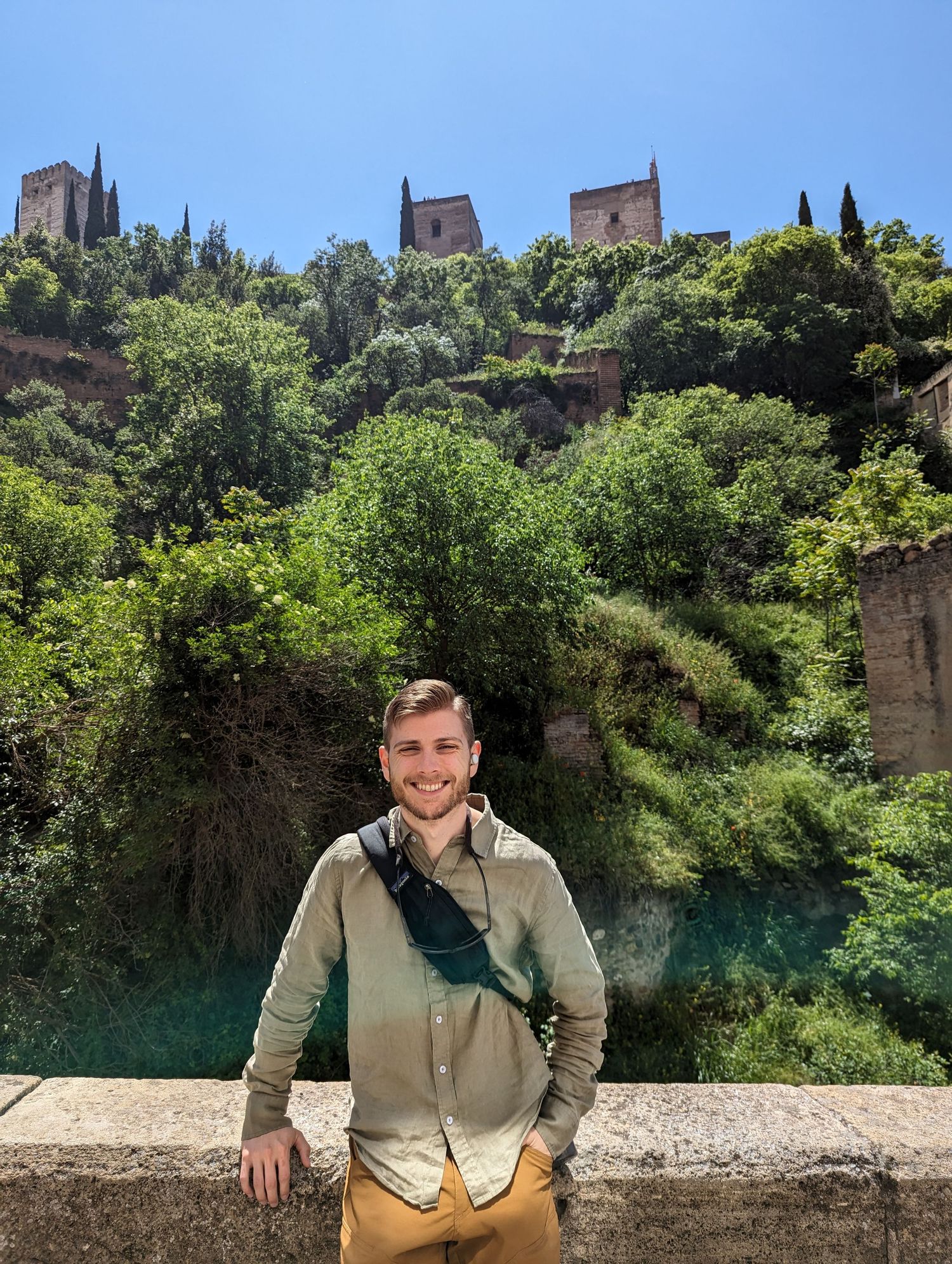 Tourist stands by a stone wall with historic architecture and lush greenery on a hillside visible in the background.