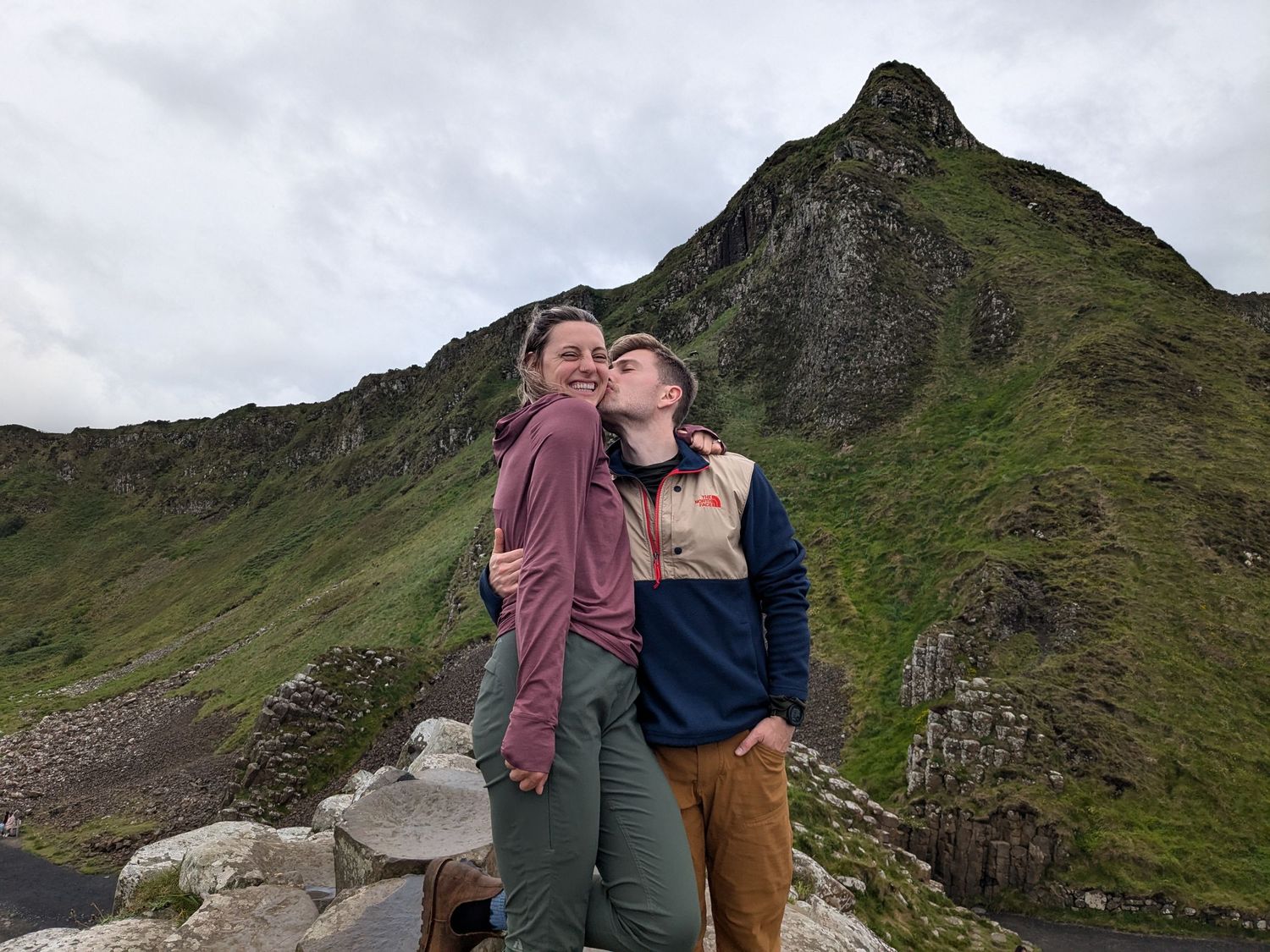 Two hikers embrace while standing on a rocky mountain path with a dramatic green peak rising behind them.