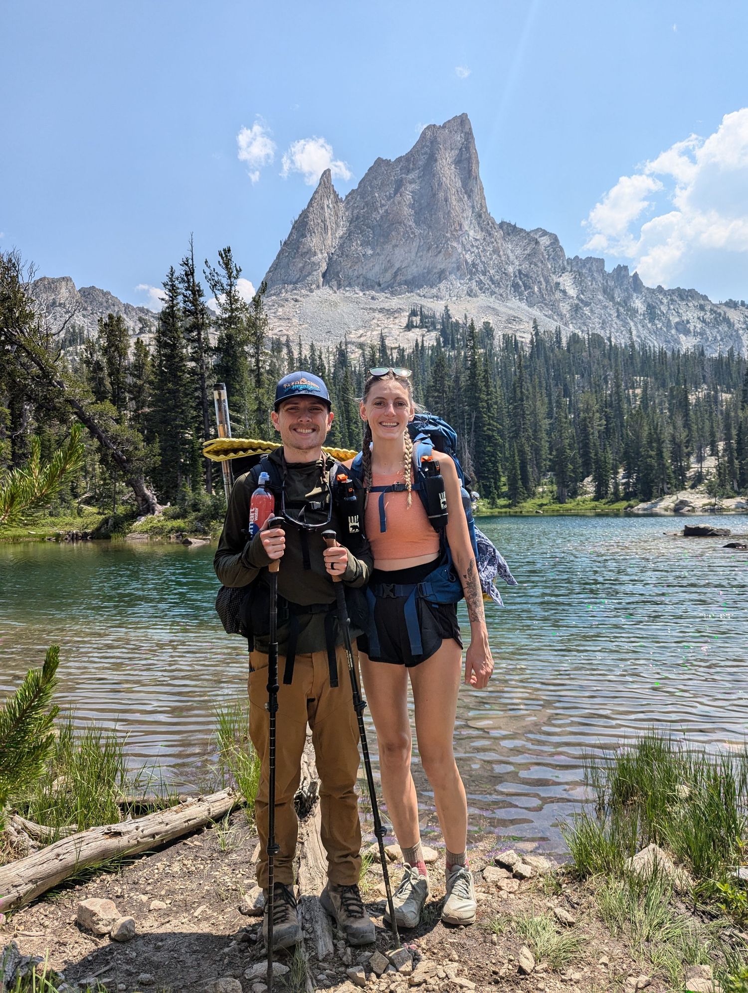 Two hikers pose at an alpine lake with dramatic mountain peak and evergreen forest backdrop in the wilderness.