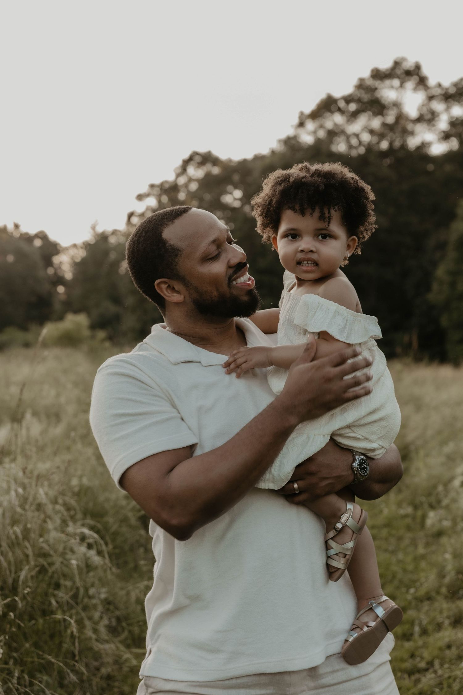 A parent in a white shirt joyfully plays with and embraces their small child in a grassy outdoor field at sunset.