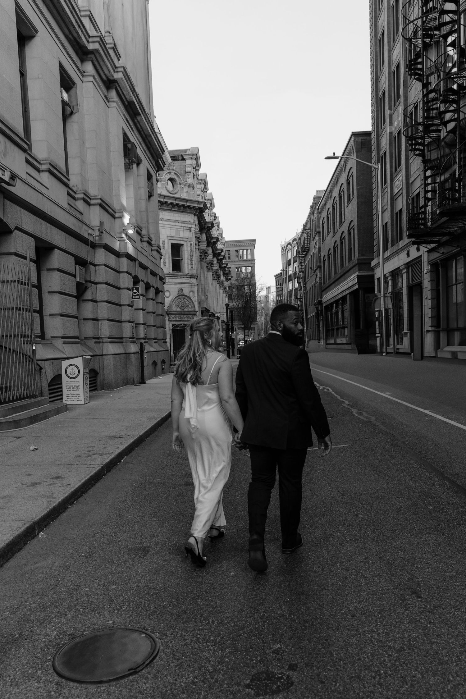 Black and white photo sequence shows two figures walking down an empty urban street with architectural buildings.