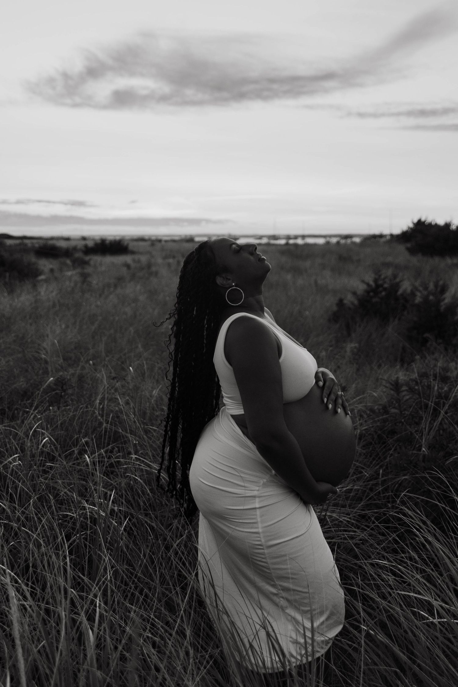 Silhouette of pregnant woman in white dress against dusky sky and tall grass.