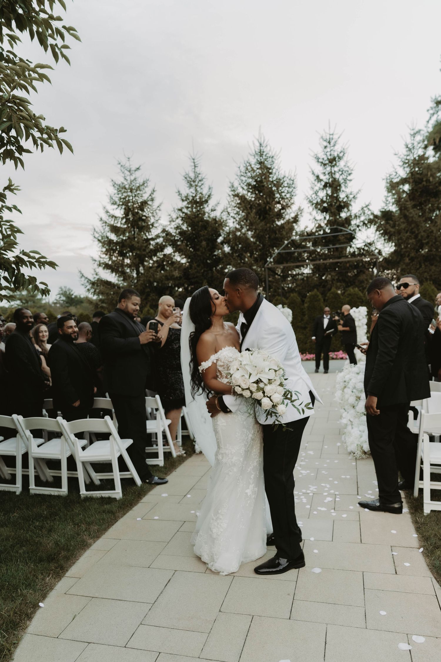 Newlyweds share a tender moment at outdoor wedding ceremony with evergreen trees and white chairs in the background.