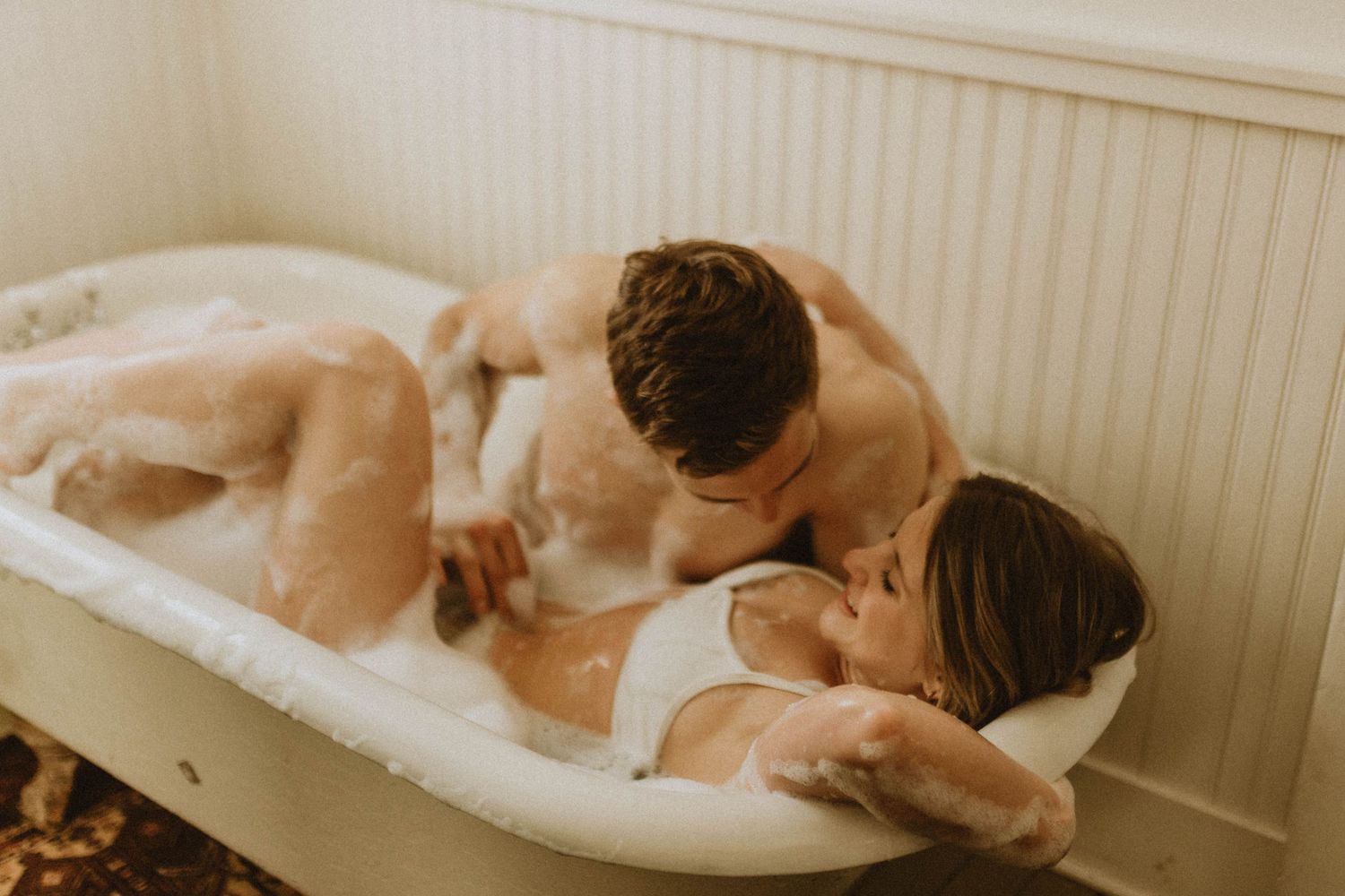Romantic couple shares tender moment in vintage clawfoot bathtub with warm lighting and soft tones.