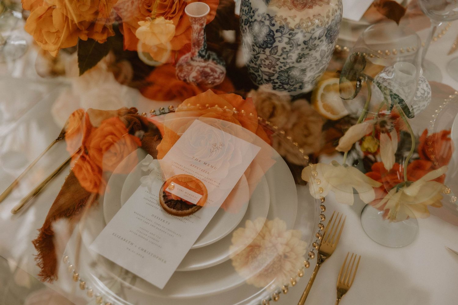Elegant wedding table setting with peach and cream florals, layered white plates, and gold utensils on a white tablecloth.