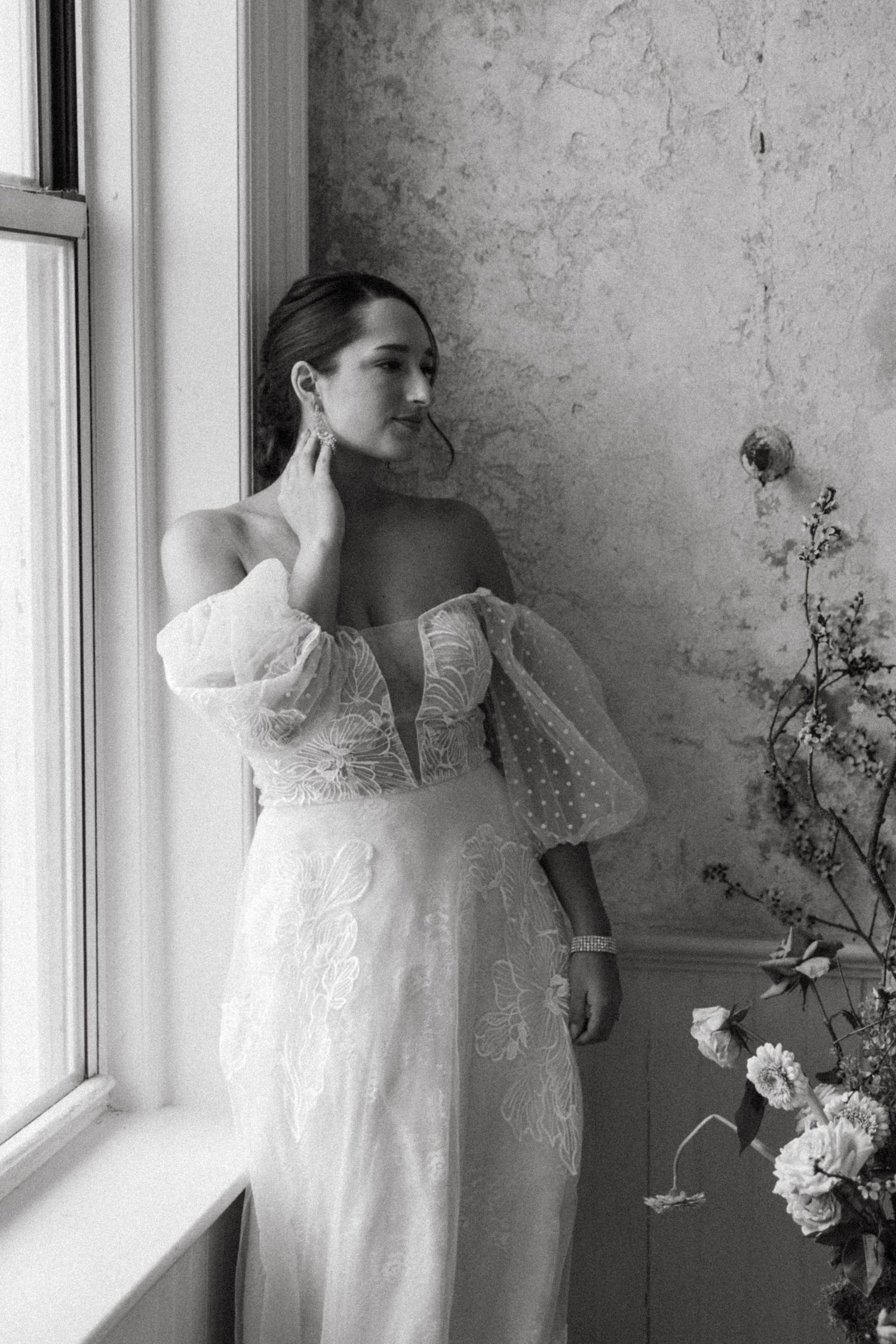 Elegant off-shoulder white lace wedding gown with puff sleeves photographed in black and white by a window.