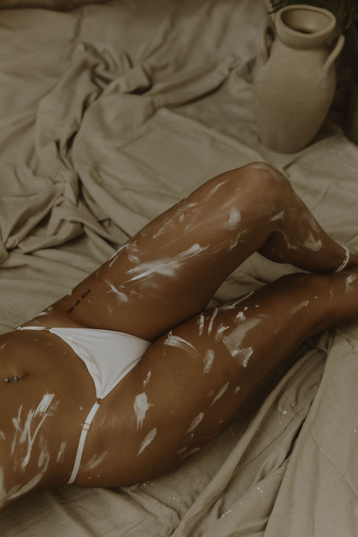 White paint splattered across a person wearing white underwear lying on beige sheets beside a ceramic bottle.