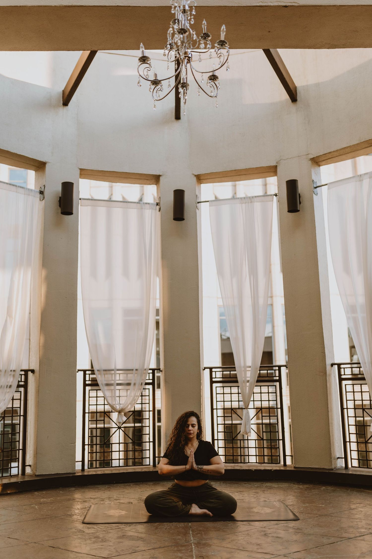 Meditative scene in sunlit room with white curtains, chandelier and person in yoga pose.