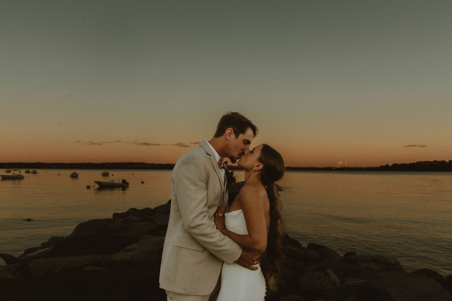 A romantic couple embraces on a rocky pier during golden hour sunset over tranquil water.