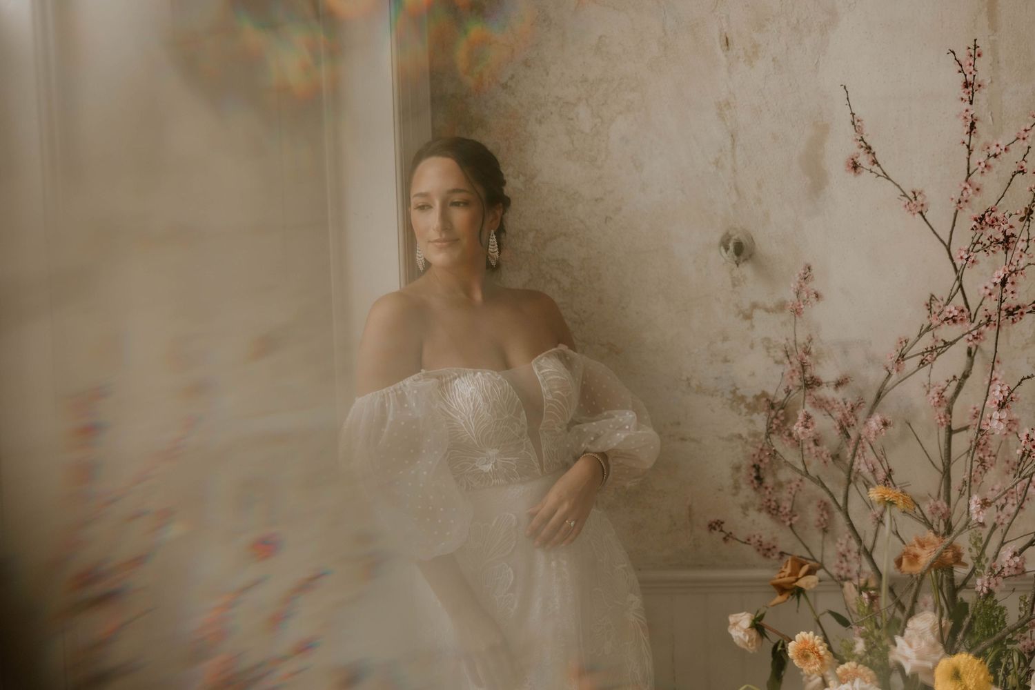 Ethereal bridal portrait in dreamy white gown with off-shoulder sleeves against textured wall with delicate floral decor.