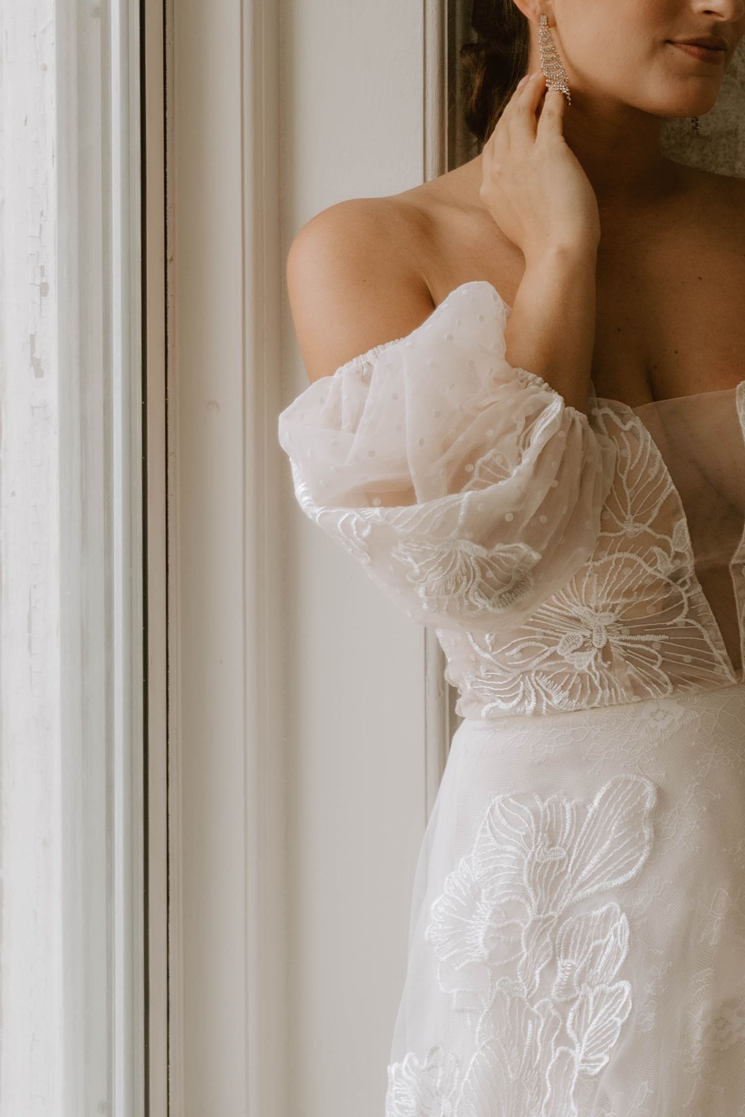 Close-up of lace wedding dress detail and off-shoulder neckline near window.