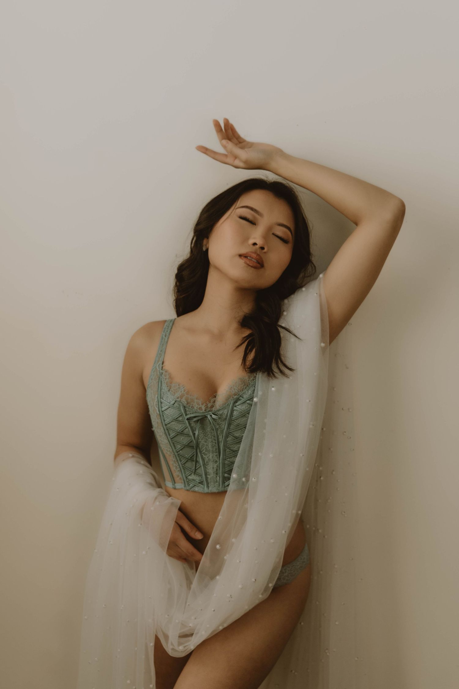 Model poses gracefully in mint green corset and sheer fabric against neutral backdrop in moody boudoir photo.