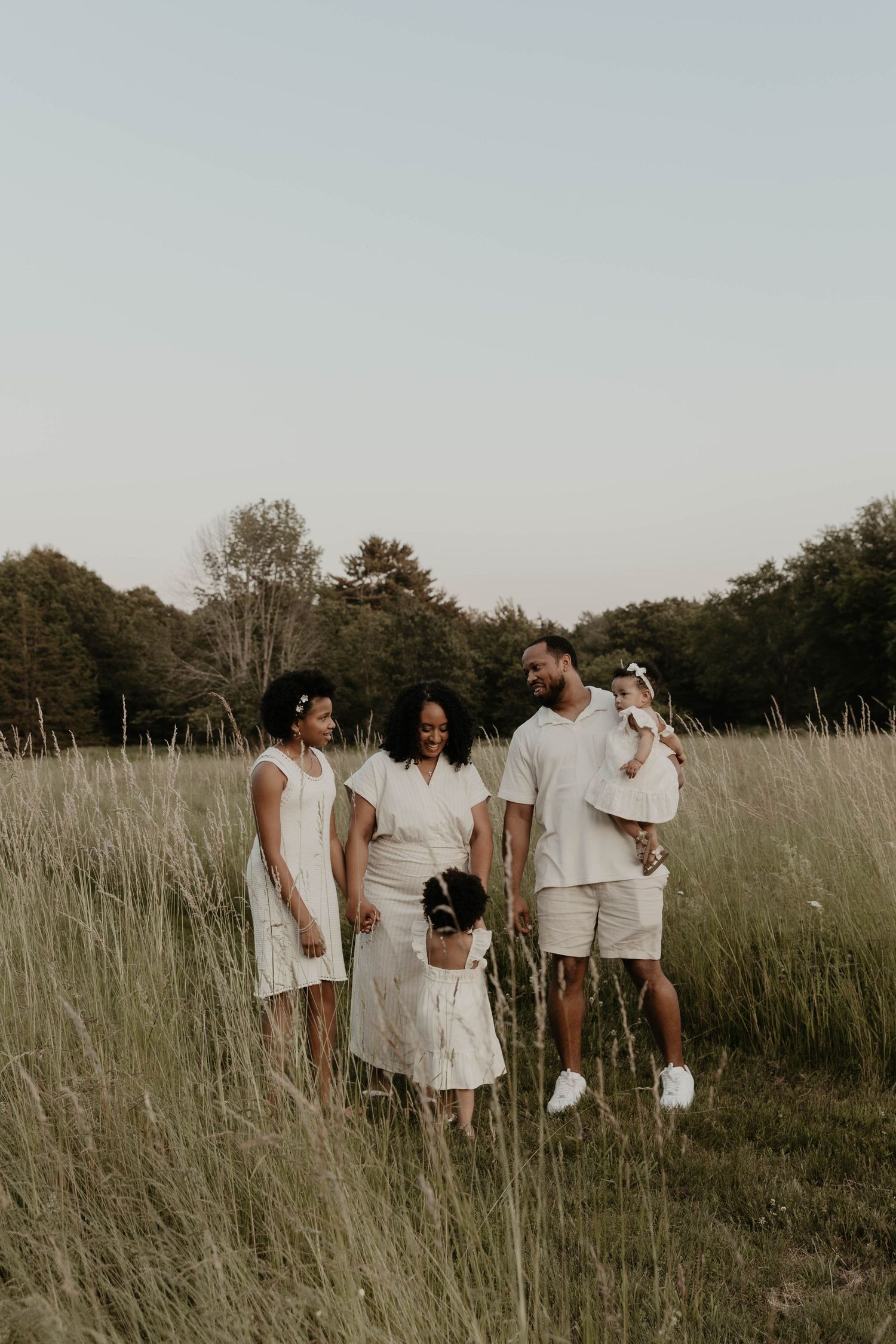 A family wearing coordinated white outfits stands together in a sunlit meadow surrounded by tall grass during golden hour.
