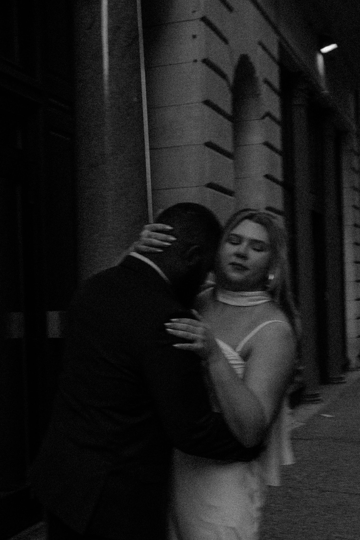 A grainy black and white photograph shows two people embracing in a dark urban alleyway at night.