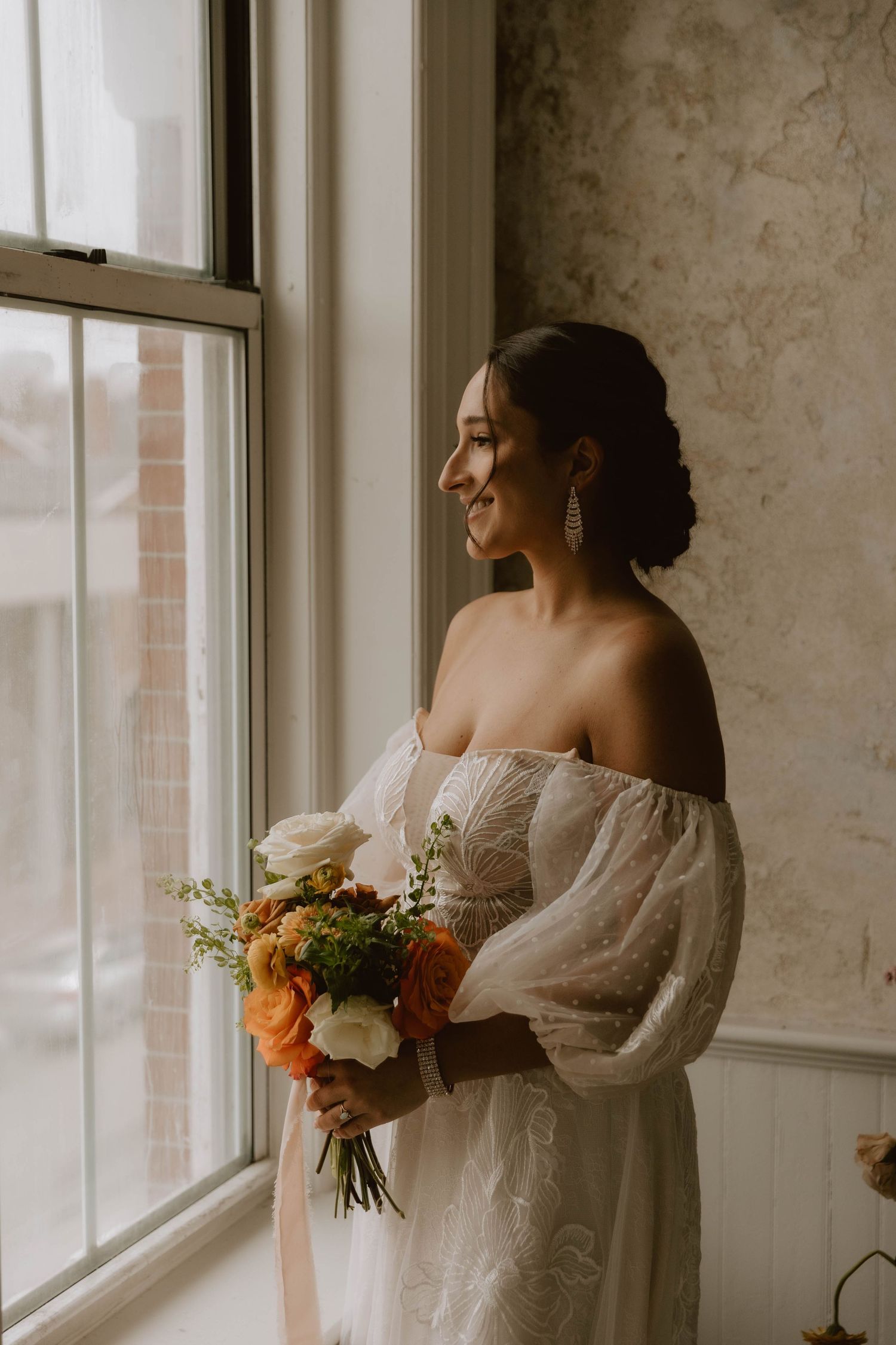 Person in off-shoulder wedding dress holding autumn bouquet by window.