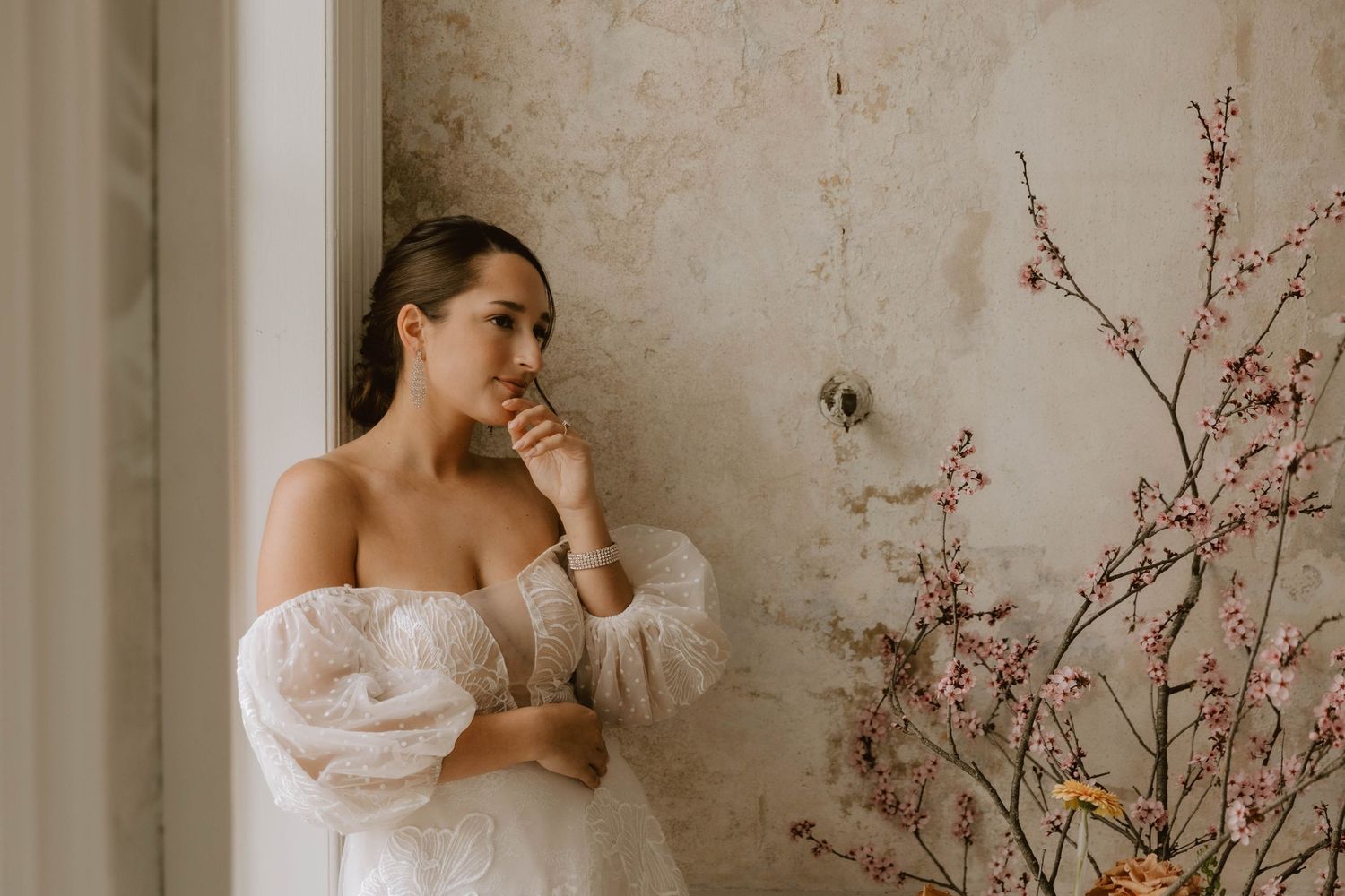 A person in a white off-shoulder dress stands thoughtfully by a window next to delicate pink cherry blossoms.