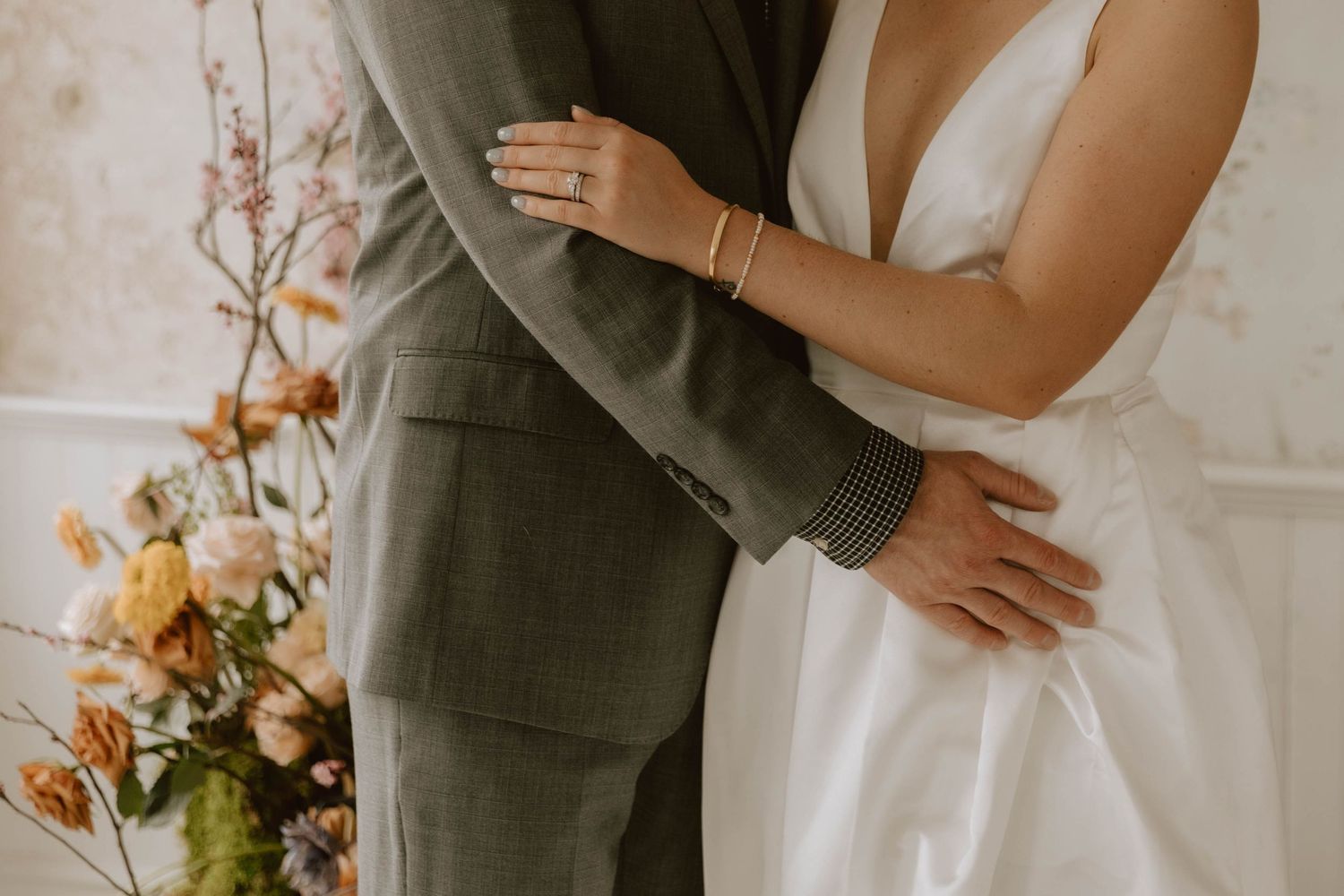 Couple embraces at wedding ceremony with bride in white dress and groom in gray suit next to autumn floral arrangement.