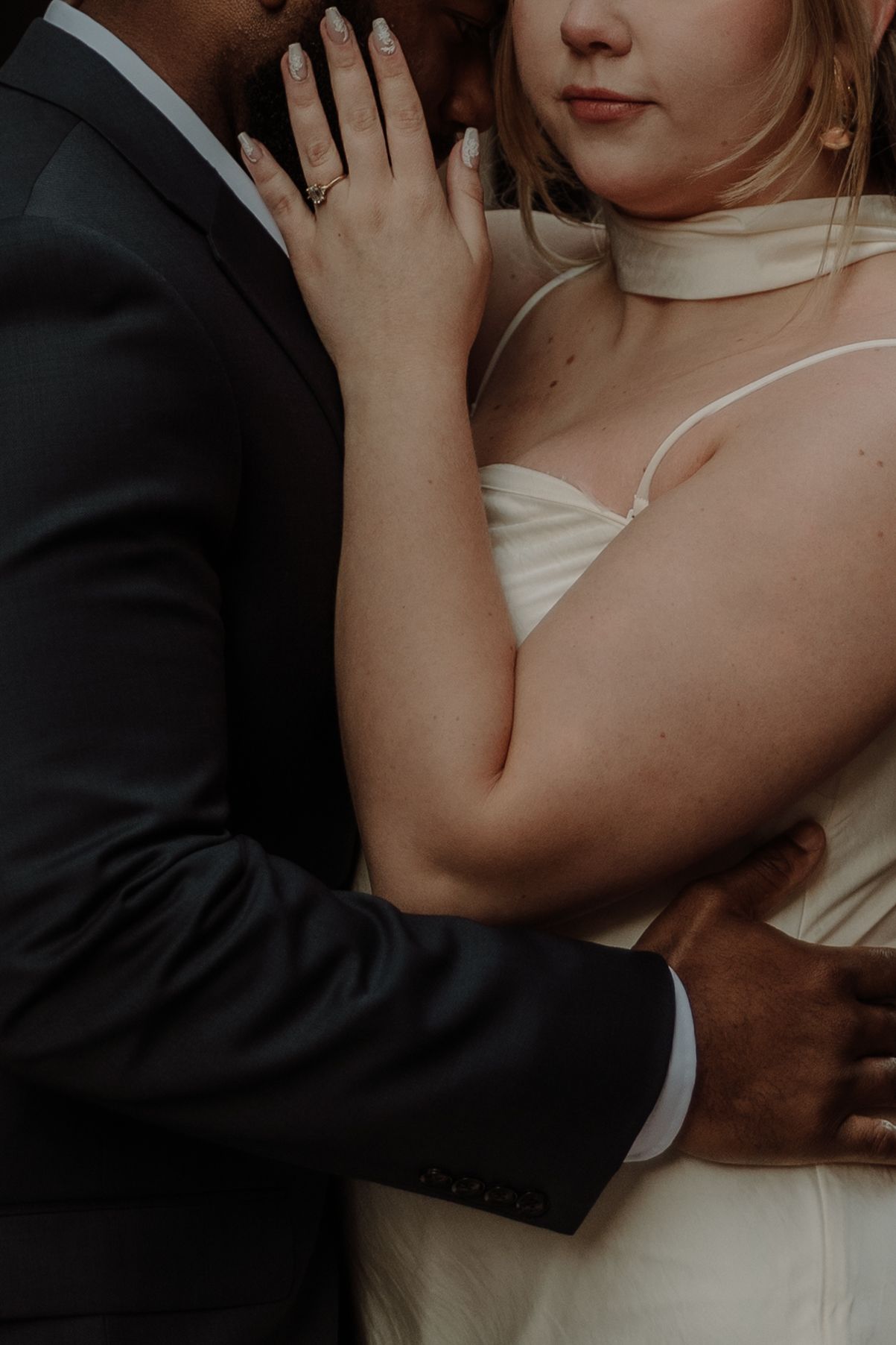 Close-up of bride and groom embracing, showing white wedding dress and black suit details in intimate moment.