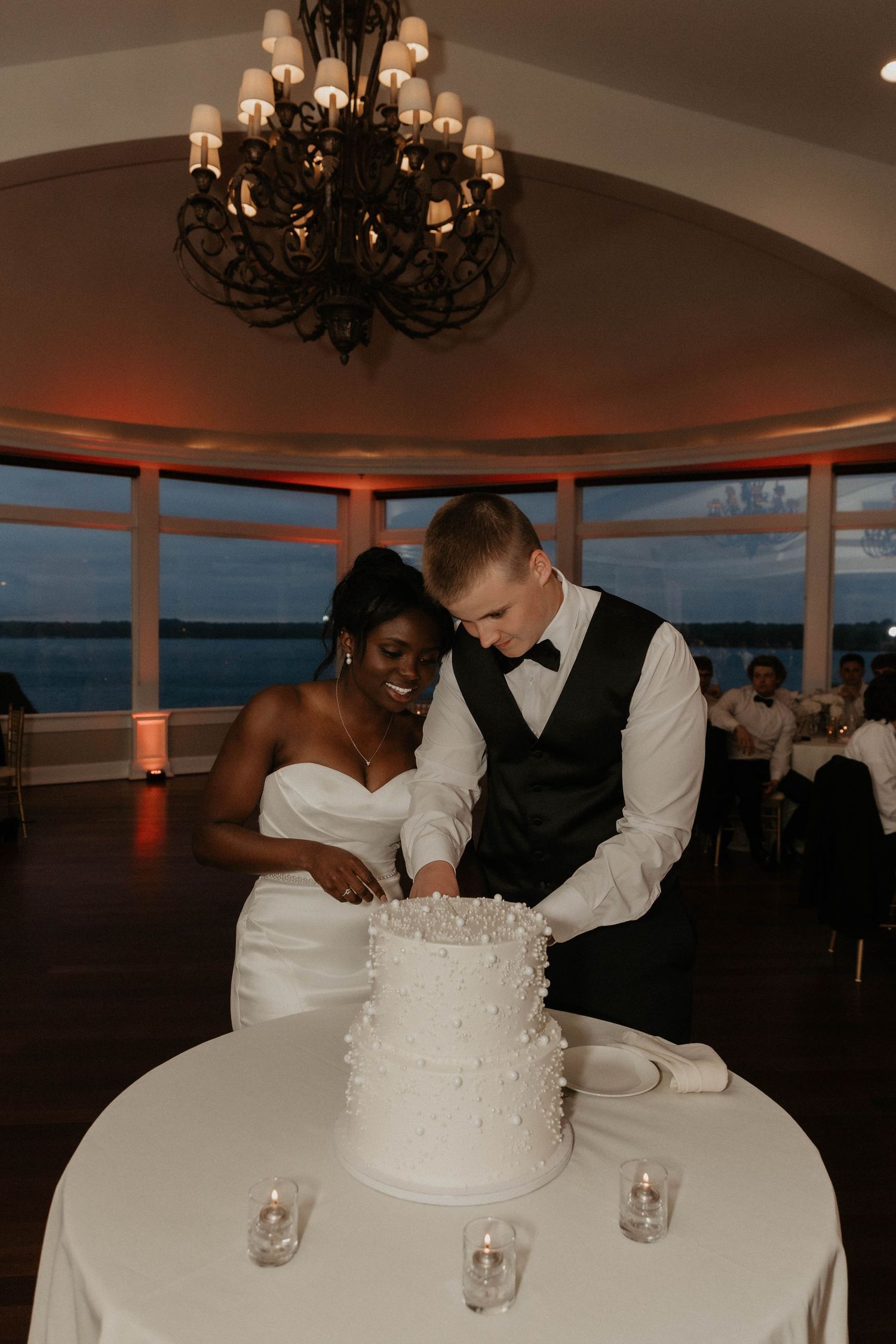 A couple cuts their elegant white wedding cake together under a grand chandelier at a waterfront reception venue.