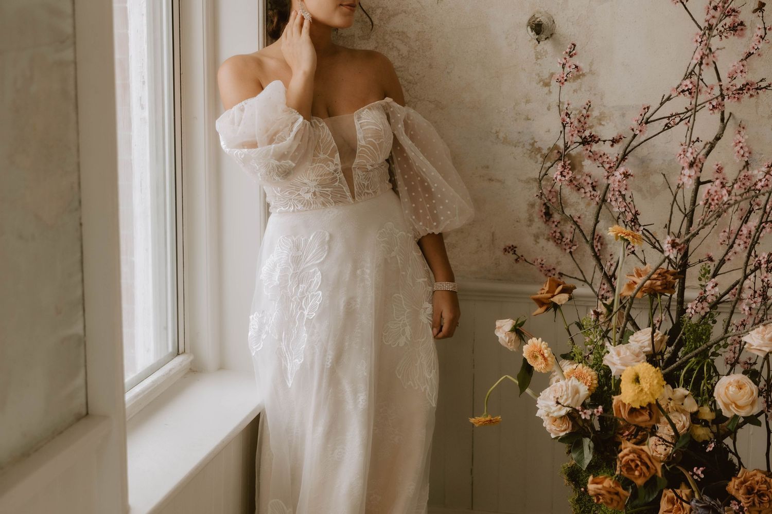 Off-shoulder white lace wedding dress with puff sleeves photographed by window next to floral arrangement.