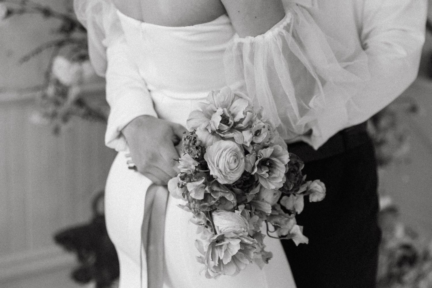 Black and white photo showing a delicate wedding bouquet with roses and mixed flowers held against a white dress.