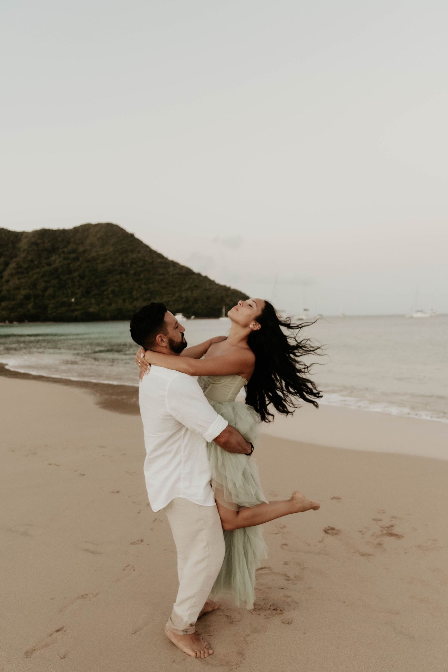 Joyful couple in white clothing embracing and laughing together on a tropical beach at sunset.