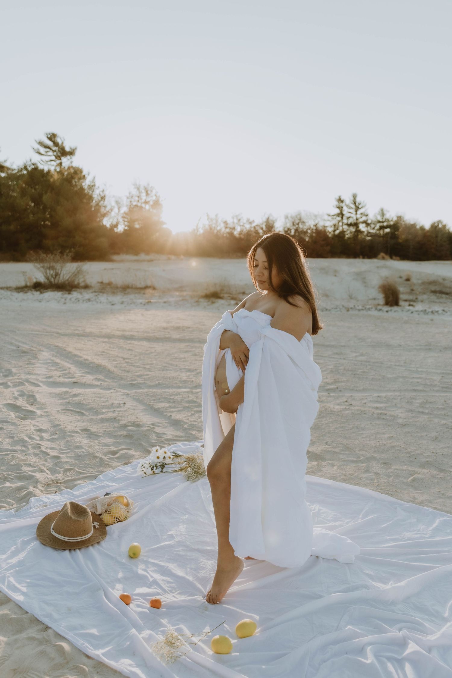 Artistic beach photoshoot with white blanket, straw hat and citrus fruits at sunset.