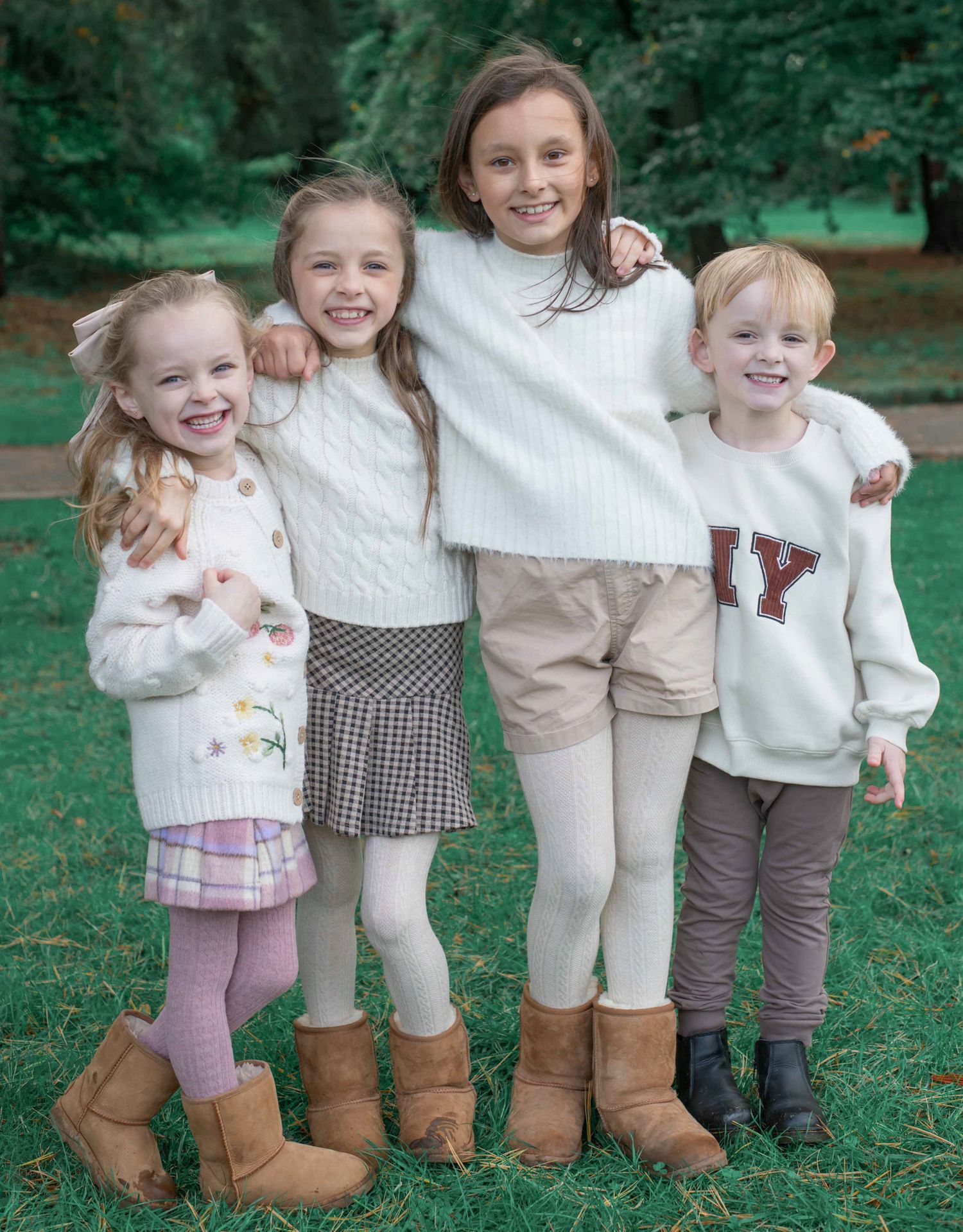 Four children wearing light colored sweaters and boots pose together on grass with trees in the background.