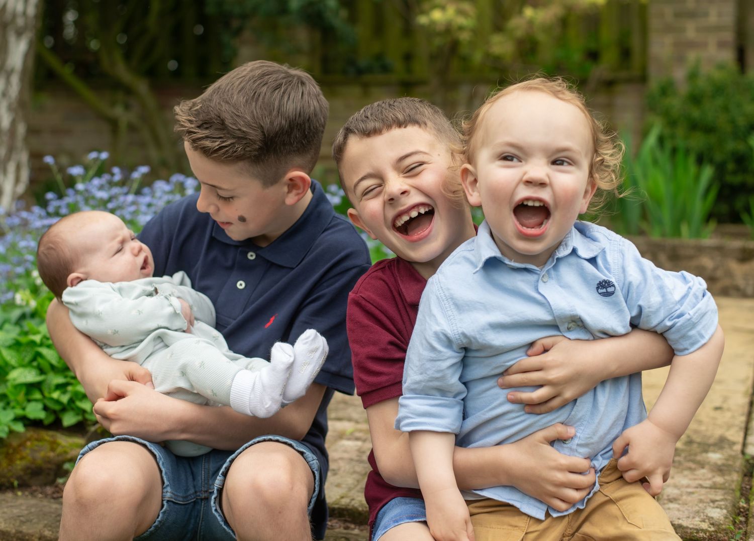 Four children sit together on a wall, with two showing big joyful laughs while one holds a baby.