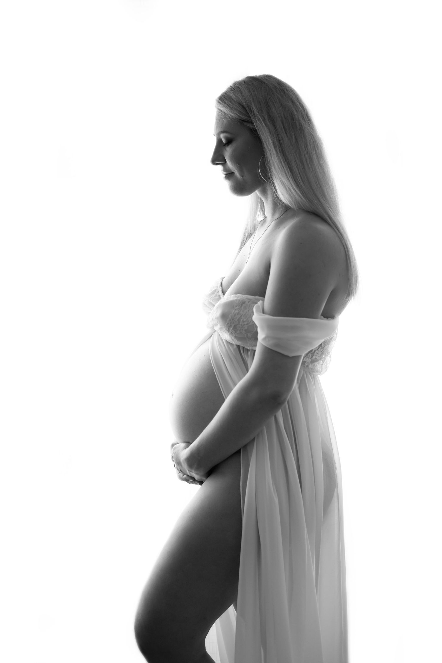 Black and white maternity photo of an expecting mother in a flowing white dress posed in profile against light background.