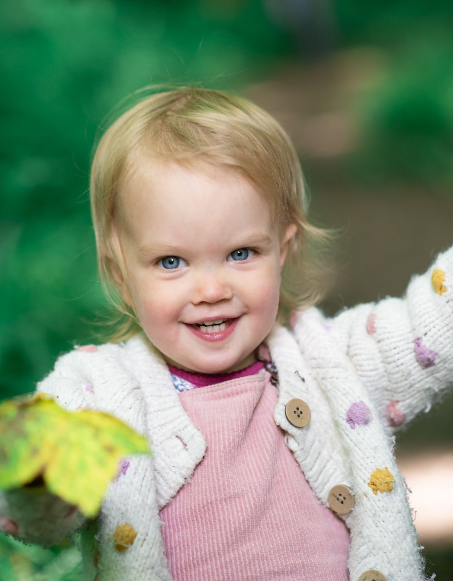 A young girl in a pink shirt and white knit cardigan smiles while standing against a green outdoor background.