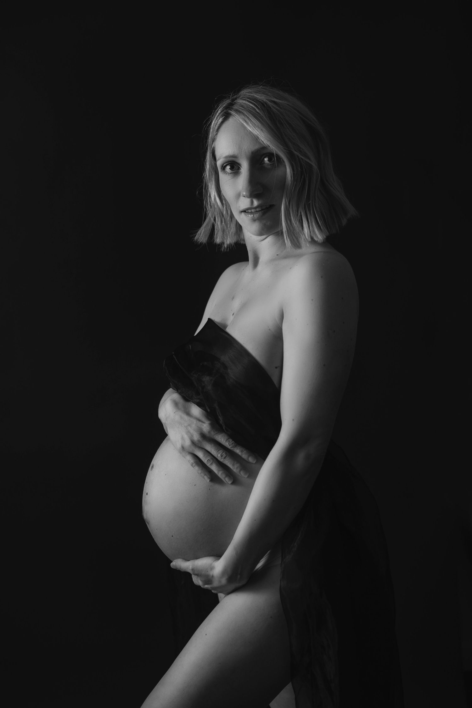 Artistic black and white maternity photo of an expectant mother in dramatic lighting with dark background.