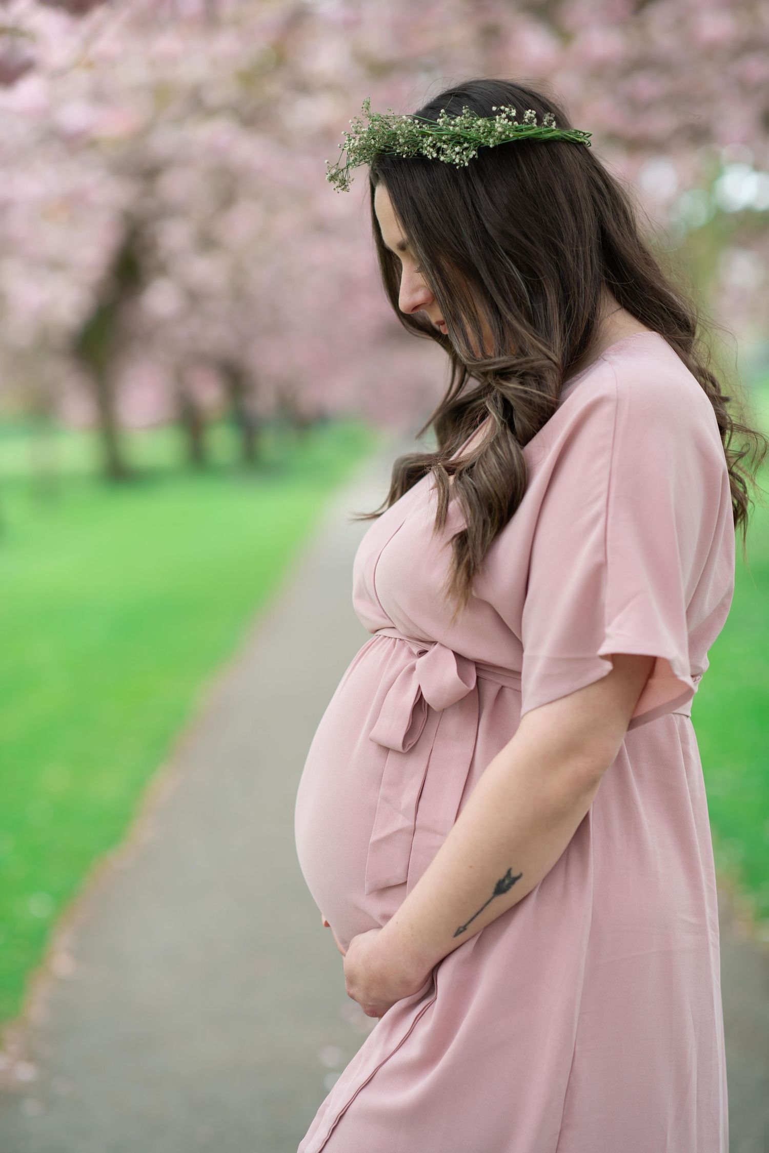 Expectant mother in pink dress and floral crown stands along cherry blossom path in spring garden.