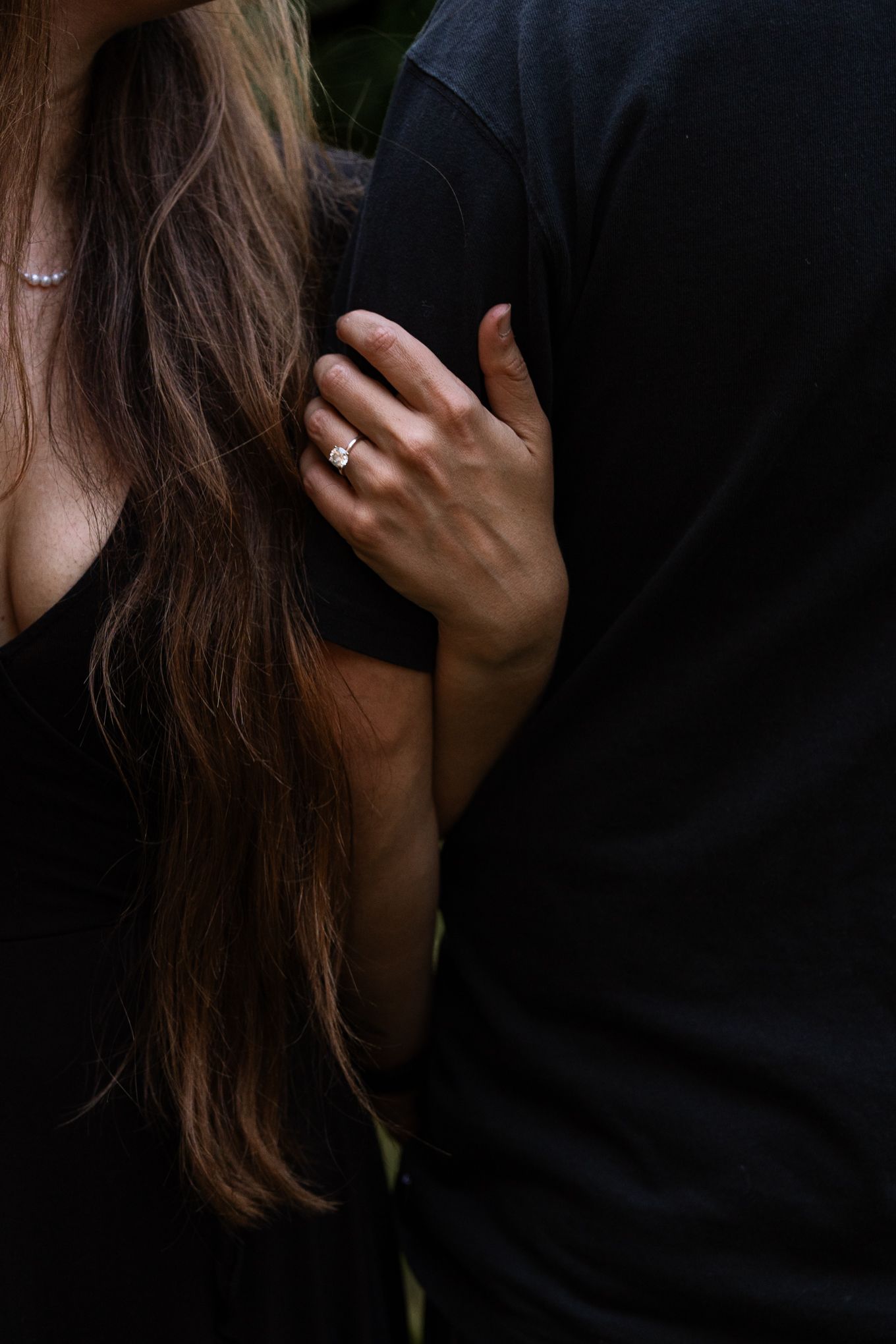 Close up detail of a diamond engagement ring on a hand against dark clothing and long brown hair.