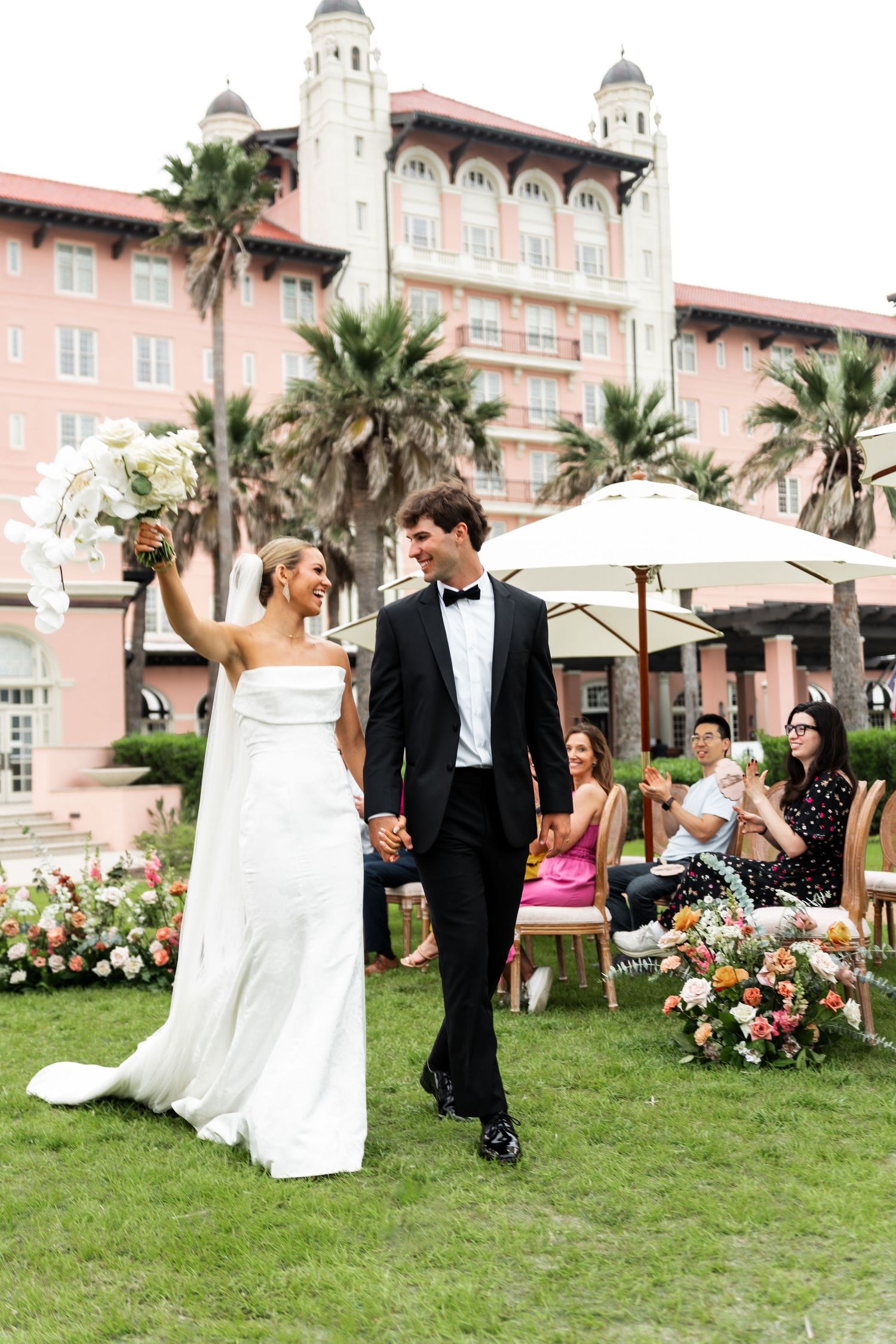 An elegant wedding ceremony takes place on a lawn in front of a historic pink hotel with Spanish architecture and palm trees.
