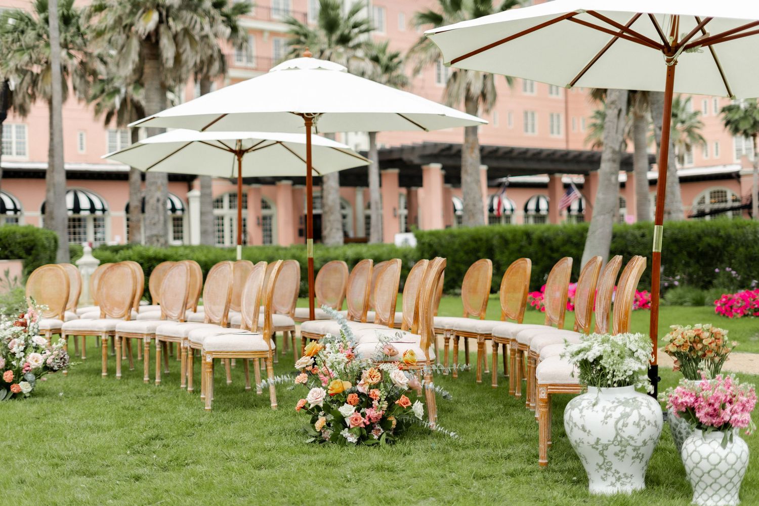 Elegant outdoor wedding ceremony setup with white chairs, floral arrangements, and umbrellas on a lawn with palm trees.