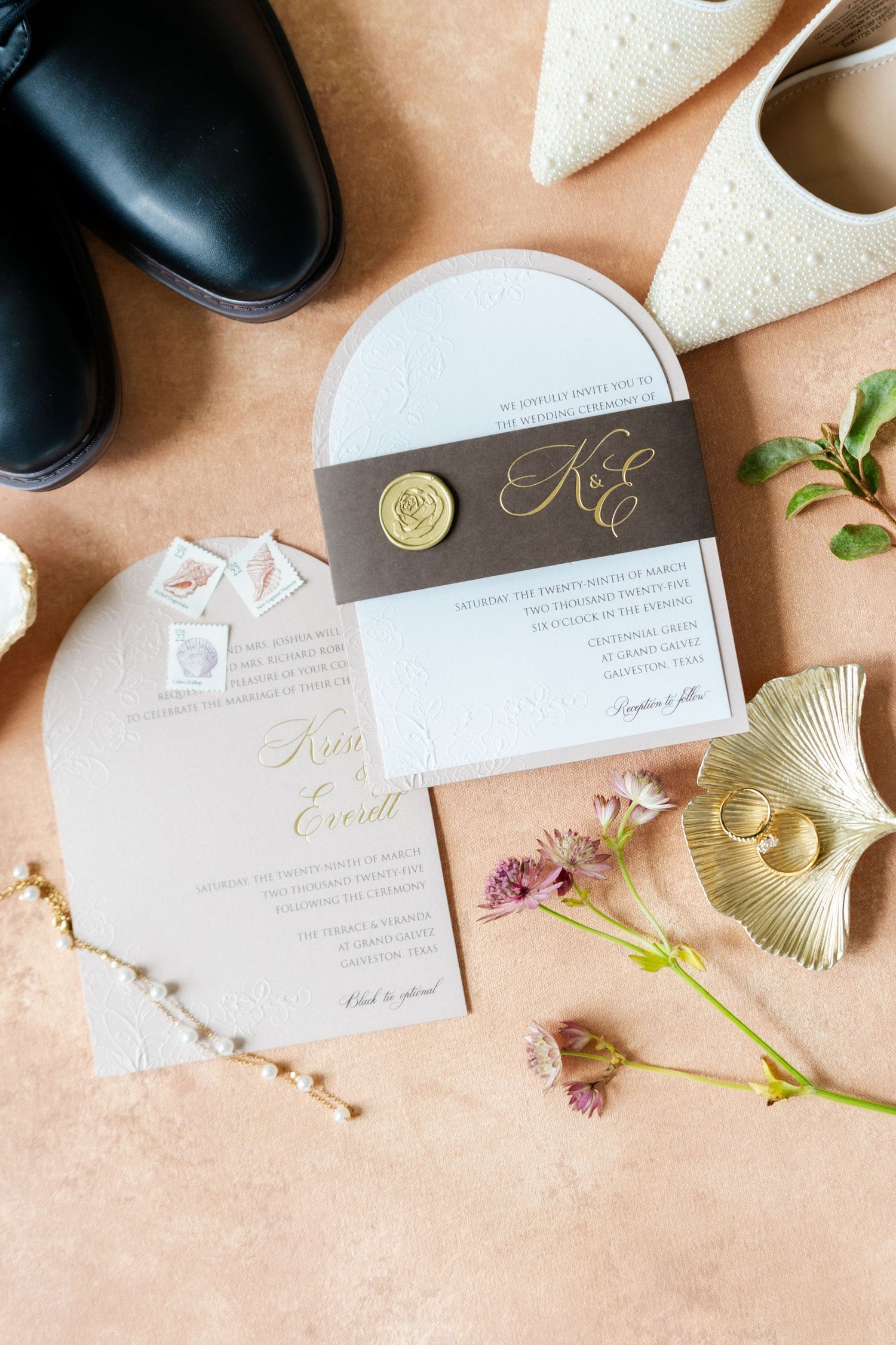 Wedding invitation suite with elegant gold lettering and wax seal displayed with dress shoes and floral accents on peach background.