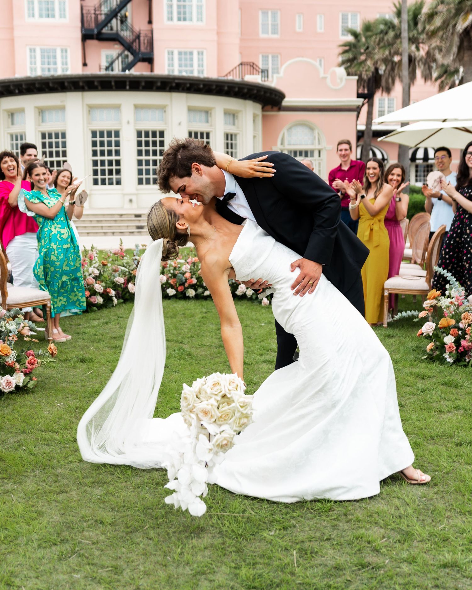 A glamorous wedding couple shares a romantic dip kiss on a lawn while guests in colorful attire watch and celebrate.