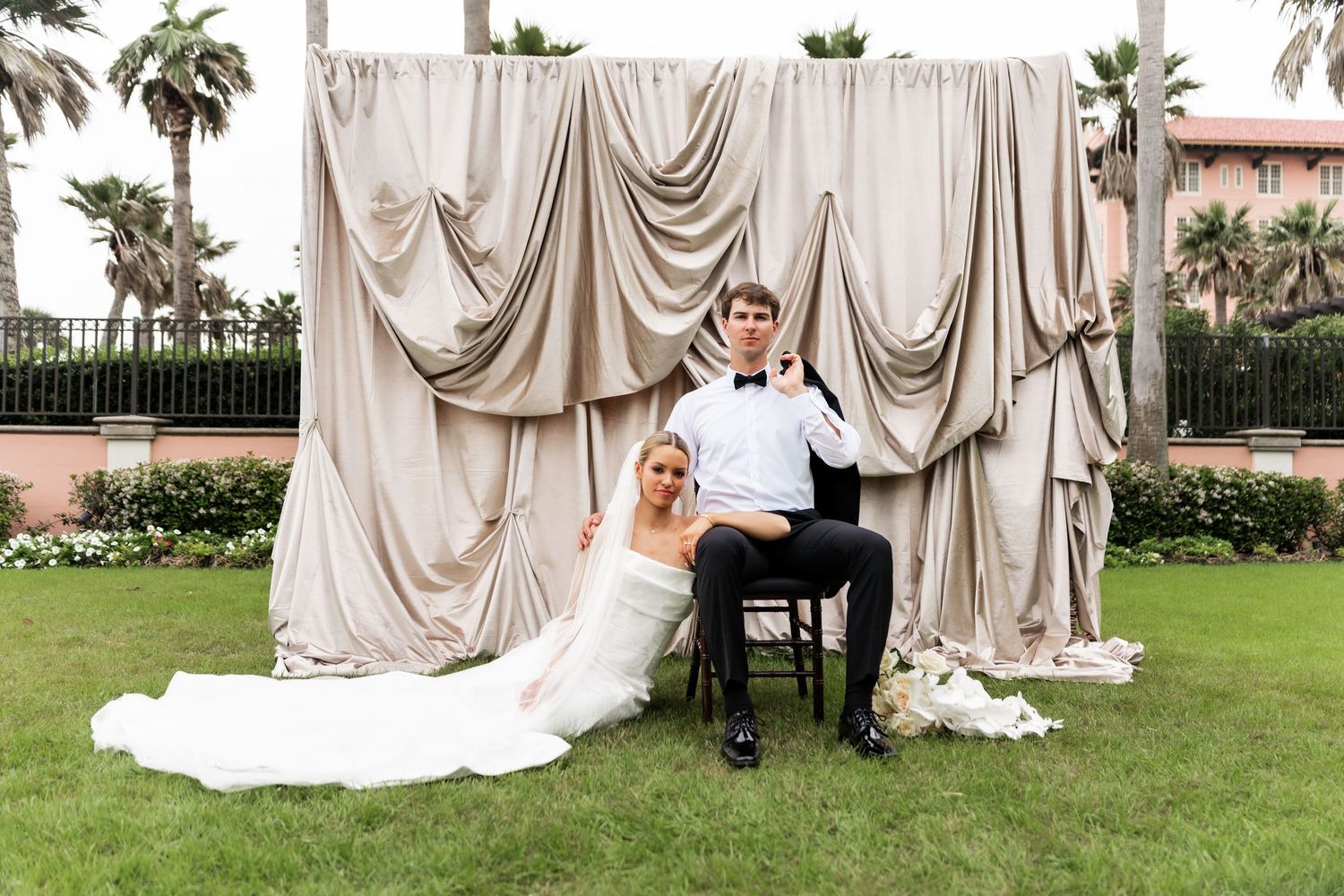 Elegant wedding ceremony on a lawn with champagne-colored draped fabric backdrop and palm trees.