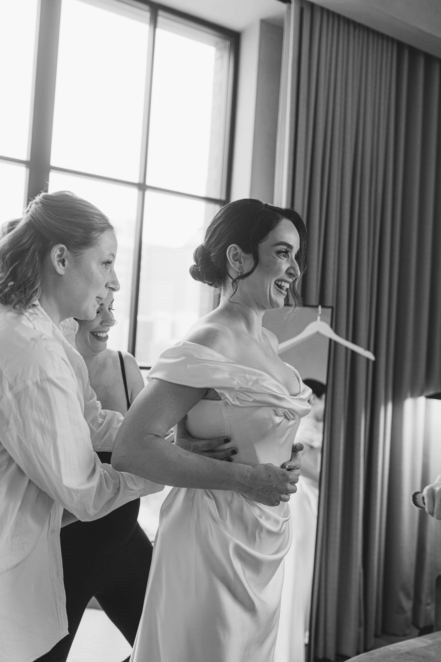 Black and white candid moment of wedding dress preparation in a sunlit room with floor-length curtains.
