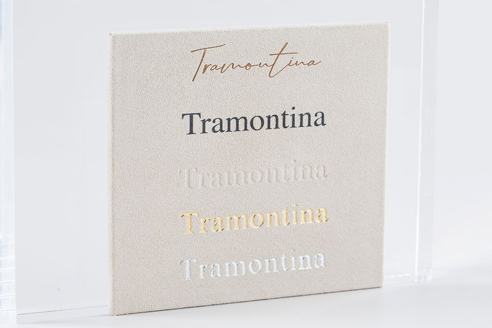 A white square ceramic tile displaying the Tramontina brand name in multiple decorative typography styles.